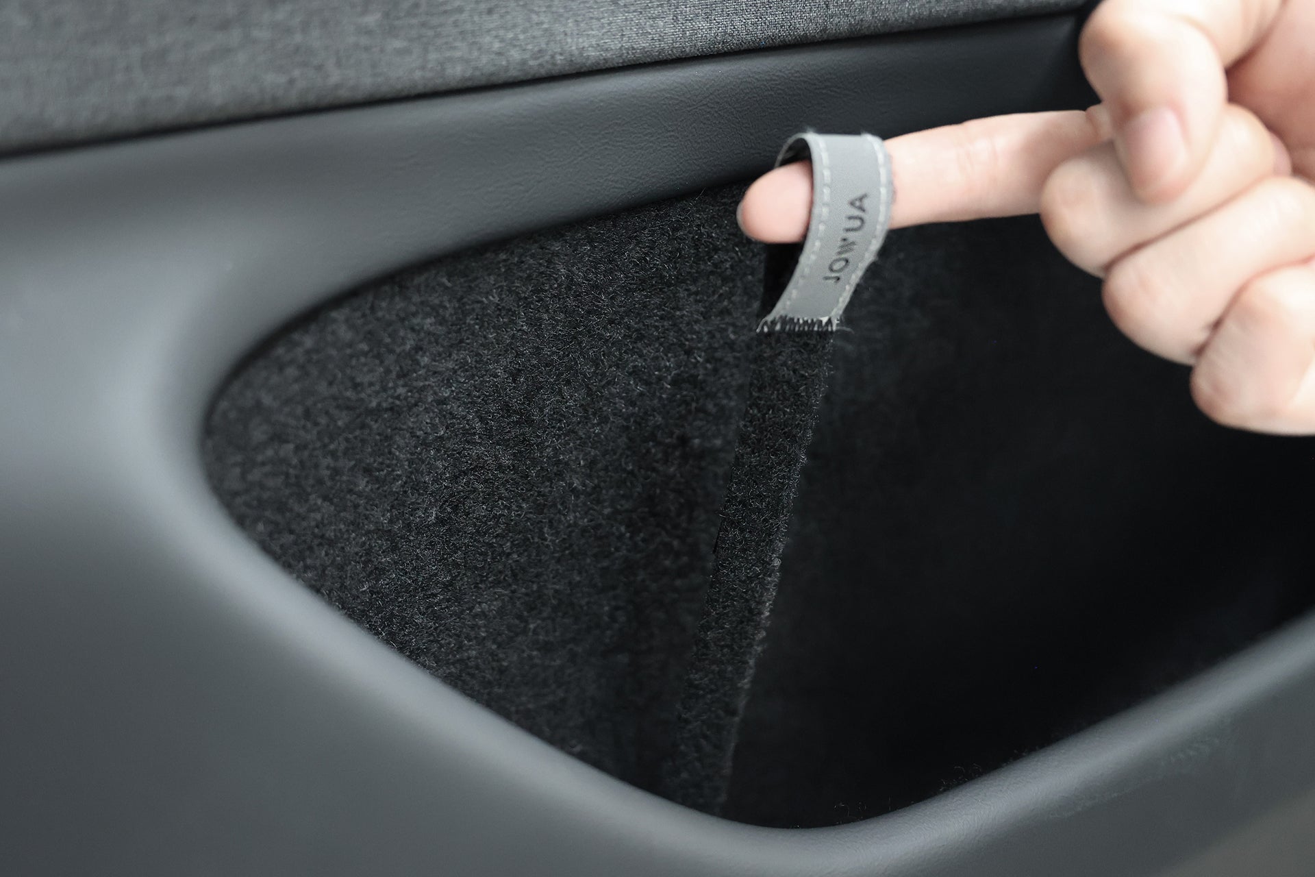 A hand pulls on the JOWUA Reflective Rear Door Emergency Release Strap, designed for Model 3 Highland (2024+) and all Model Y, attached to the inside of a car’s black, carpeted door pocket.