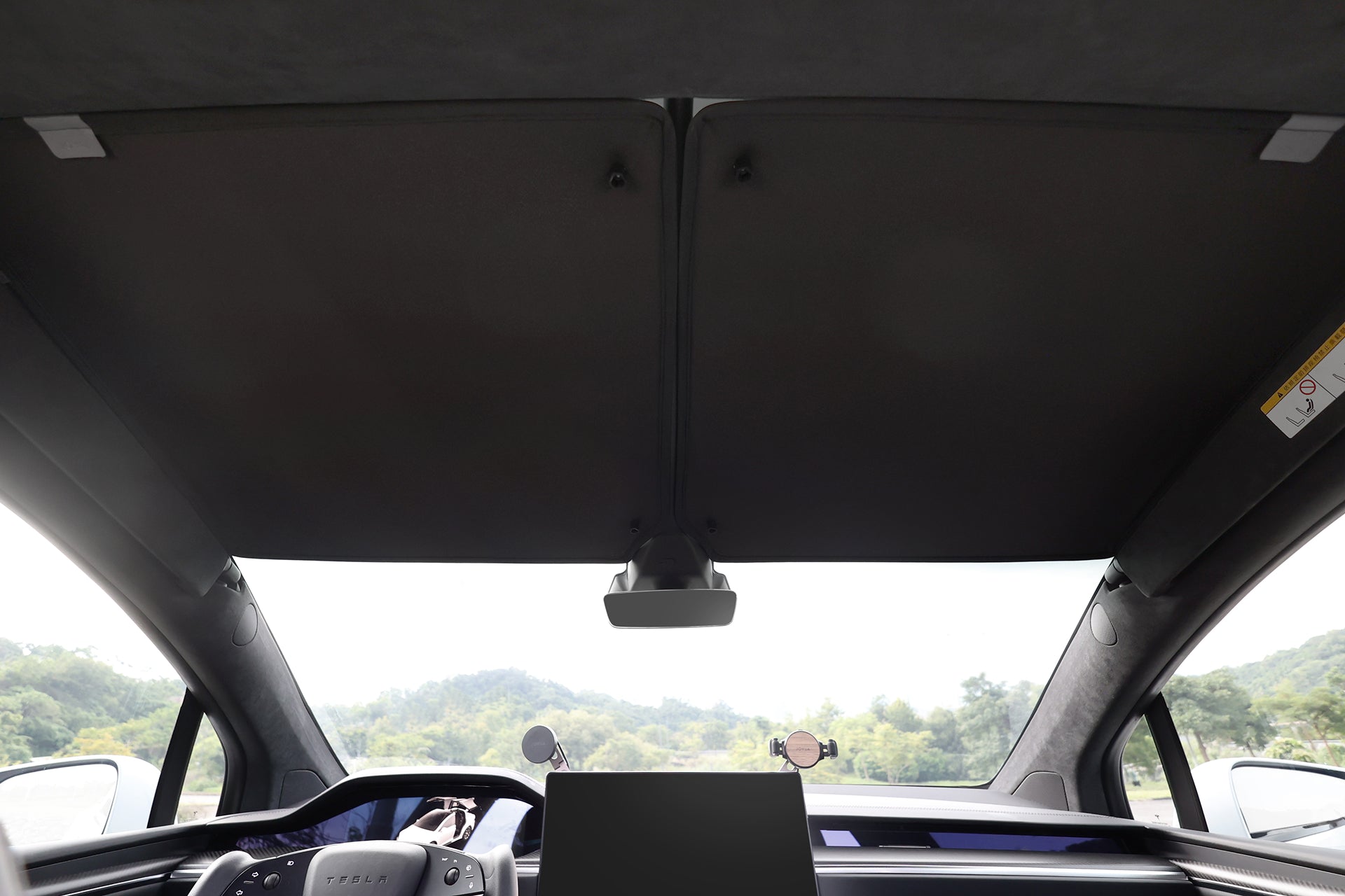 View from the back seat of a Tesla Model X featuring JOWUA's Glass Roof Sunshade (Separate Design), wide dashboard, central screen, and large UV-protective panoramic cover, with greenery visible outside.