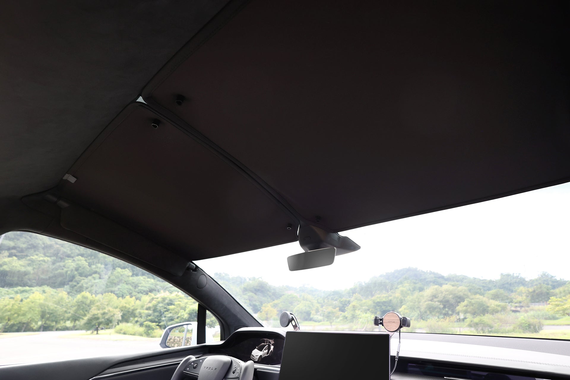 Interior view of a car with dashboard, steering wheel, large center screen, and panoramic windshield featuring the JOWUA Glass Roof Sunshade- Separate Design (Model X) for UV protection while enjoying clear views of green trees and hills outside.