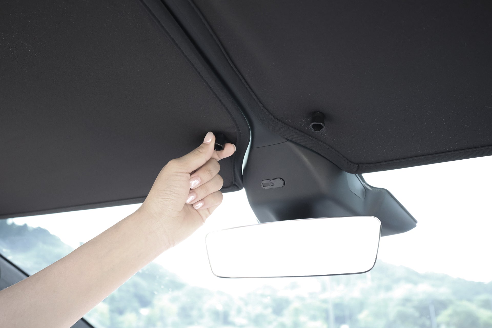 A hand adjusts the JOWUA Glass Roof Sunshade- Separate Design (Model X) near the rearview mirror, with trees visible through the windshield, enhancing UV protection and heat insulation inside.