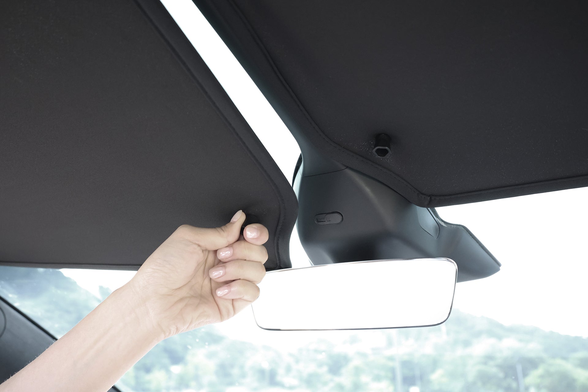A hand pulls down the JOWUA Glass Roof Sunshade- Separate Design (Model X) in a car, emphasizing UV protection and heat insulation, with the rearview mirror and blurred outdoor scenery visible through the windshield.