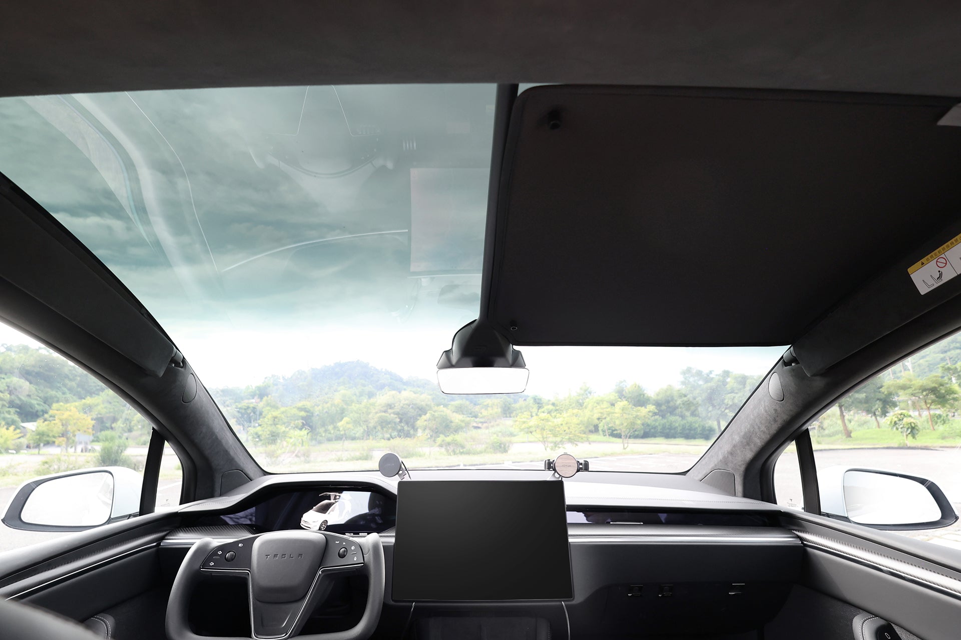 Interior view of a car with a large windshield, modern dashboard, central touchscreen, and side mirrors. The JOWUA Glass Roof Sunshade- Separate Design (Model X) provides UV protection, with trees and greenery visible outside the windows.