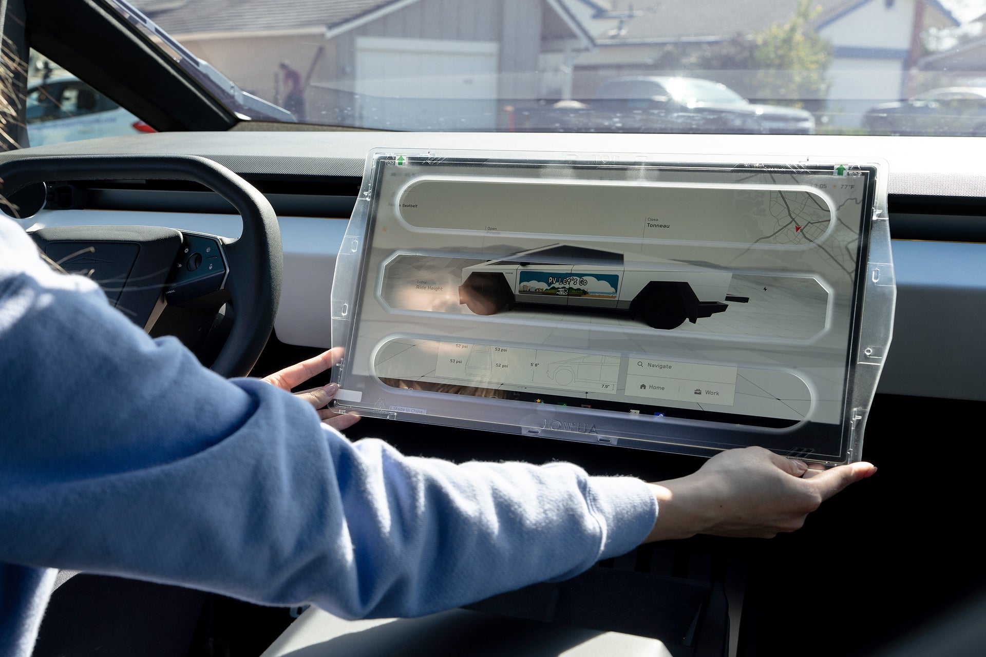 A person in a blue sleeve holds the JOWUA Tempered Glass Screen Protector for Cybertruck, displaying a vehicle interface inside a modern car with houses visible through the windshield in the background.