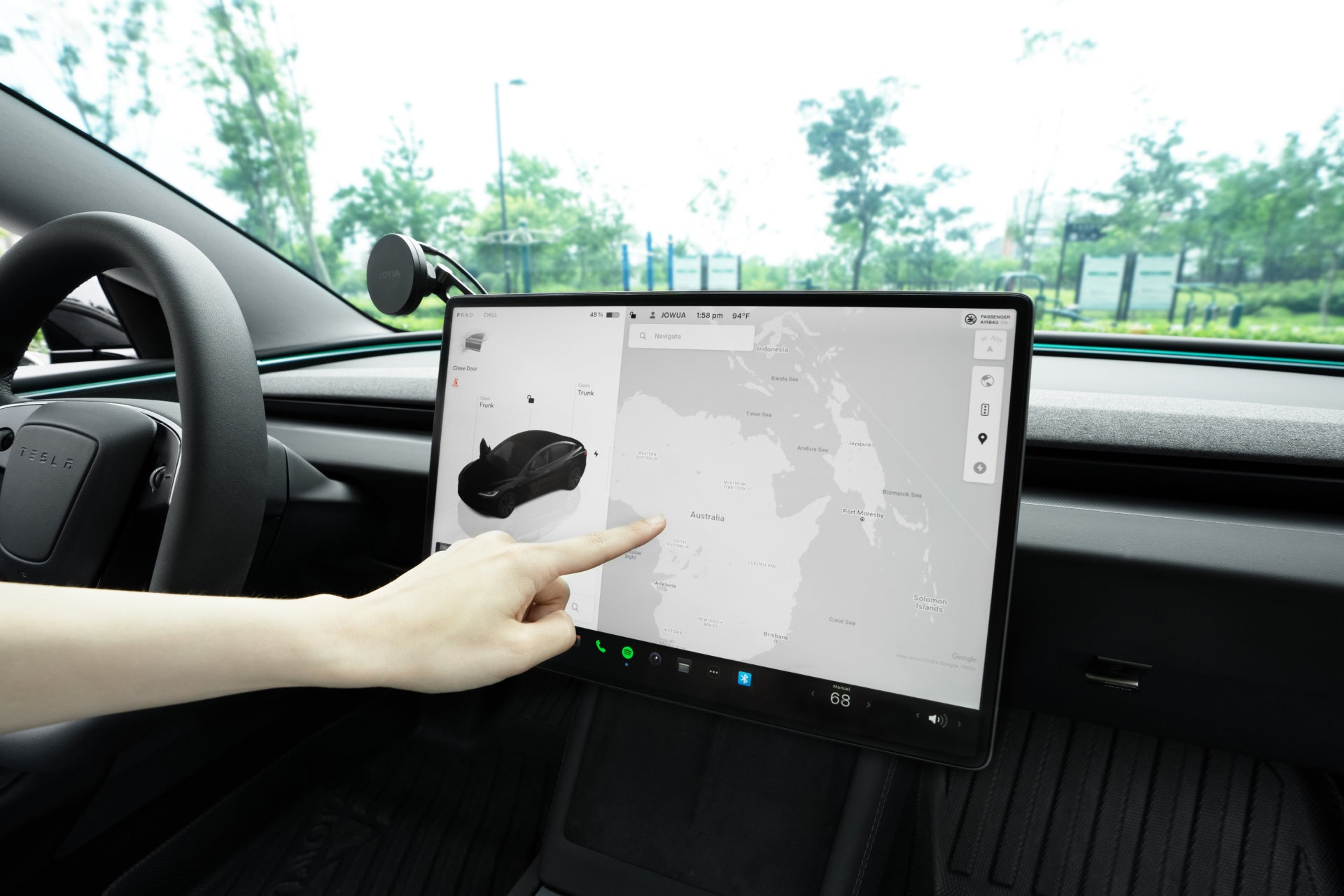 A person’s hand points at the large touchscreen of a Tesla Model Y 2025+ or Model 3 2024+, fitted with a JOWUA Tempered Glass Screen Protector (15"/16"), displaying navigation and controls, with trees and signs visible through the windshield.