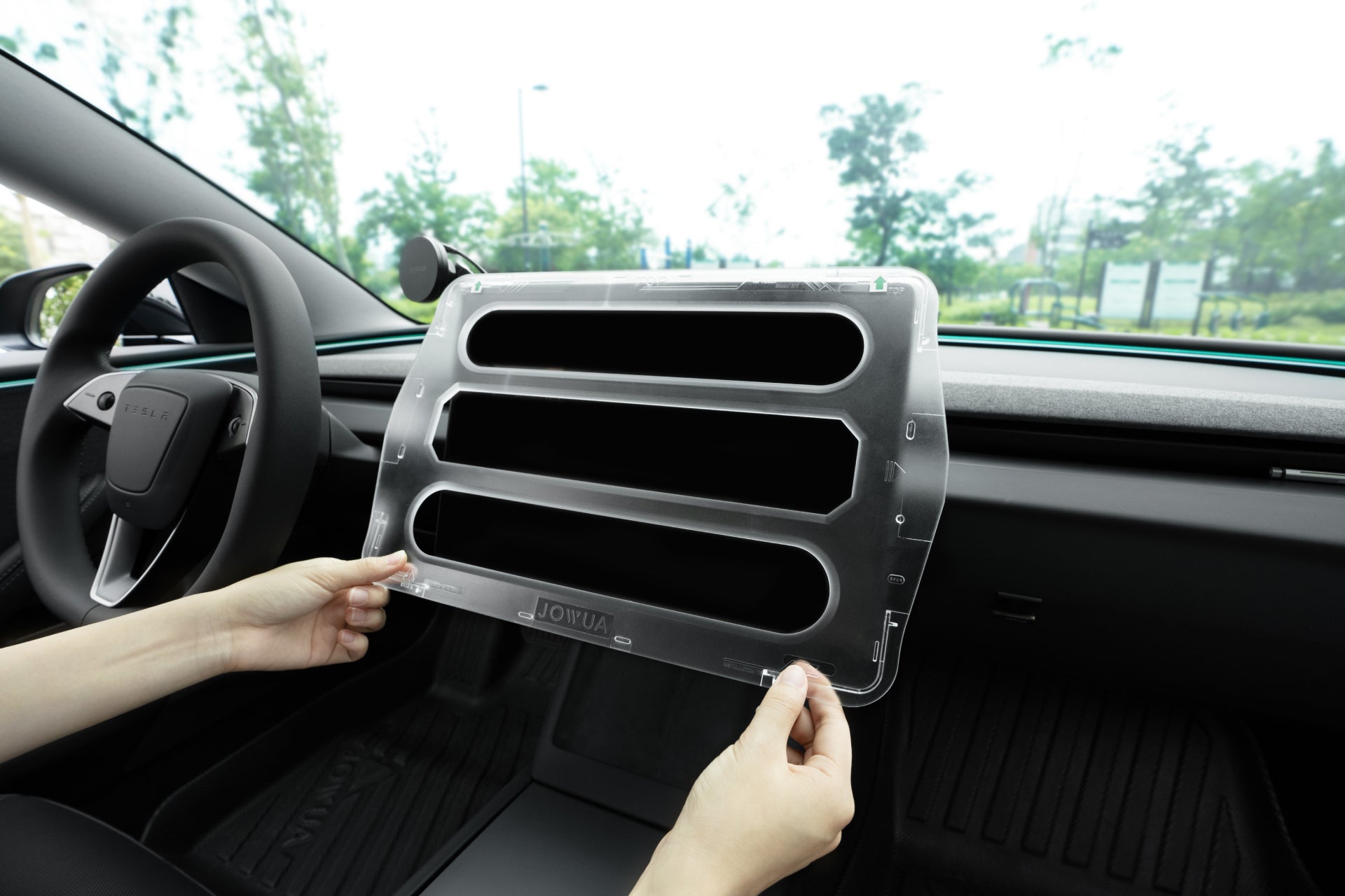 Inside a JOWUA Tesla Model Y Juniper 2025+ Premium Bundle (RHD), someone holds a transparent plastic component with three horizontal slots resembling a vent cover. The clean, modern interior complements the design, while trees are visible through the windshield.