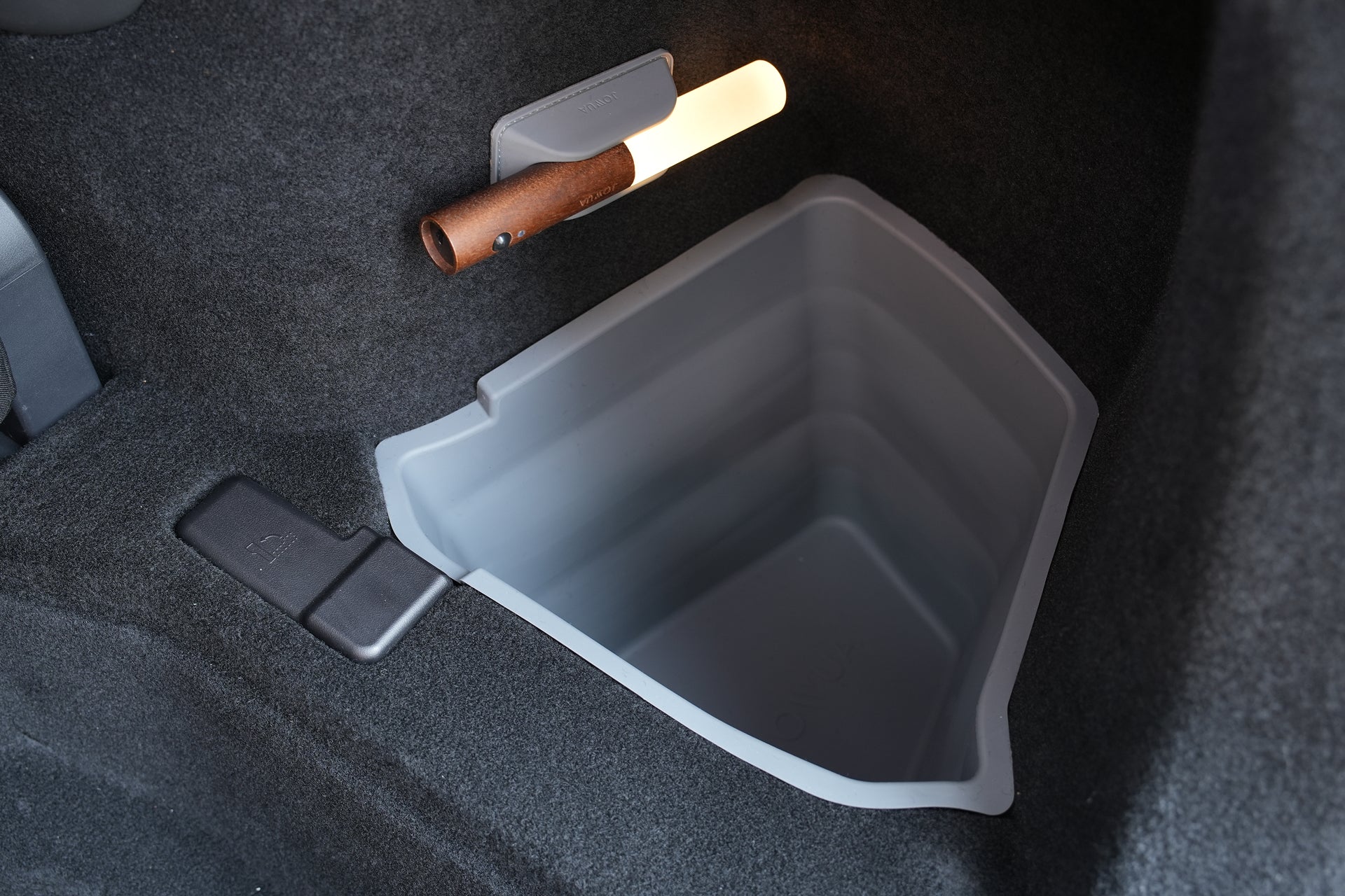 A gray, deep JOWUA Tesla Model Y L (2026+) trunk storage bin is integrated into the carpeted floor of the Model Y L’s trunk. Docked above the bin, a cylindrical flashlight with a brown handle is securely held in its dedicated mount. The silicone organizer features an angular, geometric design and is part of the Trunk Storage Bins + Ambient Light Bundle.