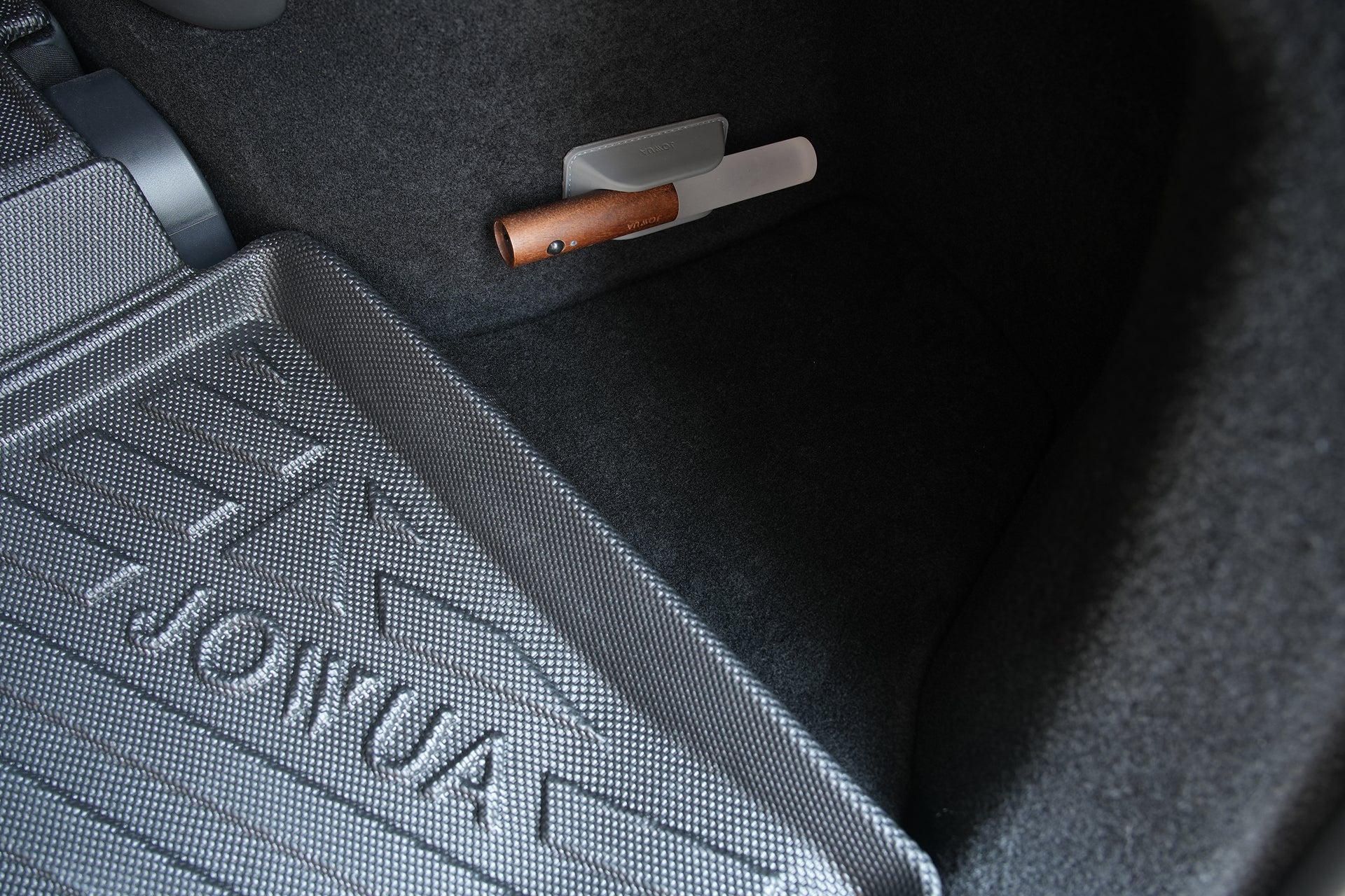 A close-up view of a car's floor mat with a textured surface and the word "JOWUA." A small wooden-handled brush rests in a holder attached to the side carpeting, next to JOWUA's Tesla Model Y L (2026+) Trunk Storage Bins + Ambient Light Bundle, designed specifically for your Tesla Model Y L trunk.