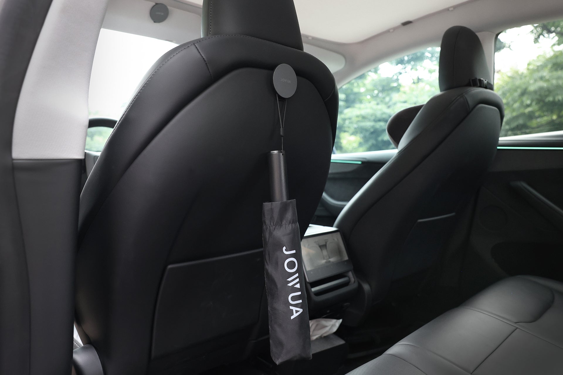 A JOWUA Type-C Electric Umbrella in black hangs from the back of a front car seat inside a modern vehicle with black leather interior, with trees visible through the window.