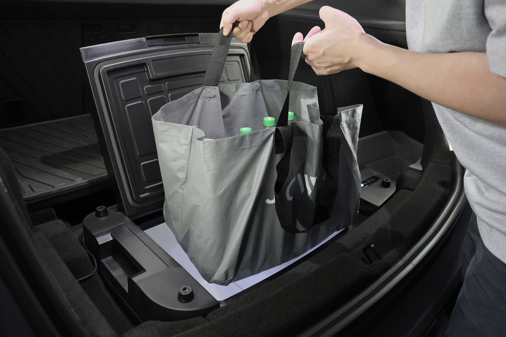 A person places a gray reusable shopping bag with green-capped bottles into the open front trunk of a Tesla Model Y equipped with the JOWUA Car Refrigerator. The person's arms are visible, and the intelligent dual-mode cooling system is featured.