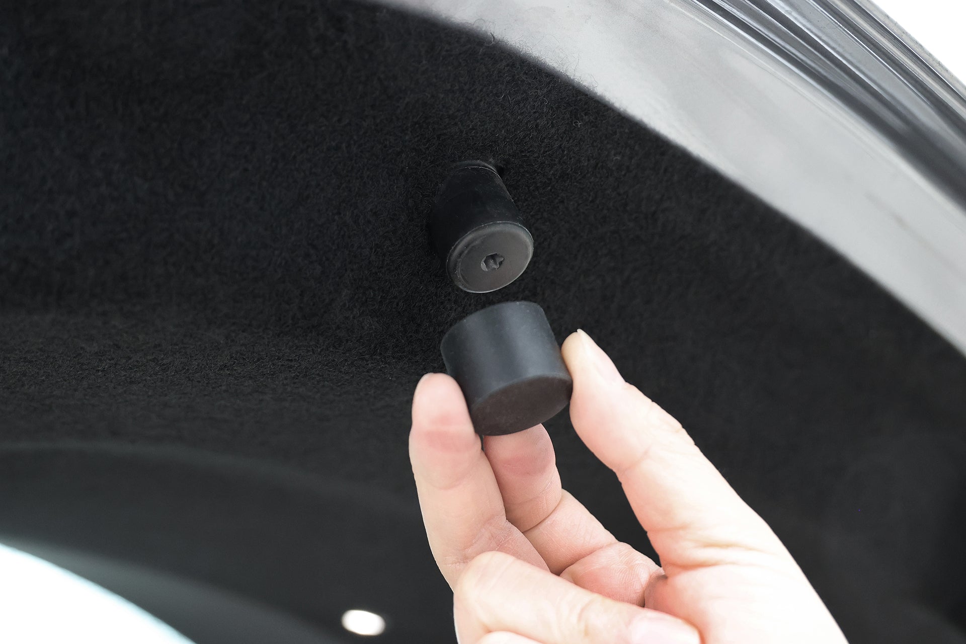 A hand holds a small black rubber stopper near a similar stopper on the inside of a Tesla Model Y trunk lid, showing where the JOWUA Teslussy Light > Type-C Ambient Light & 3 in 1 Car Charger can be installed.