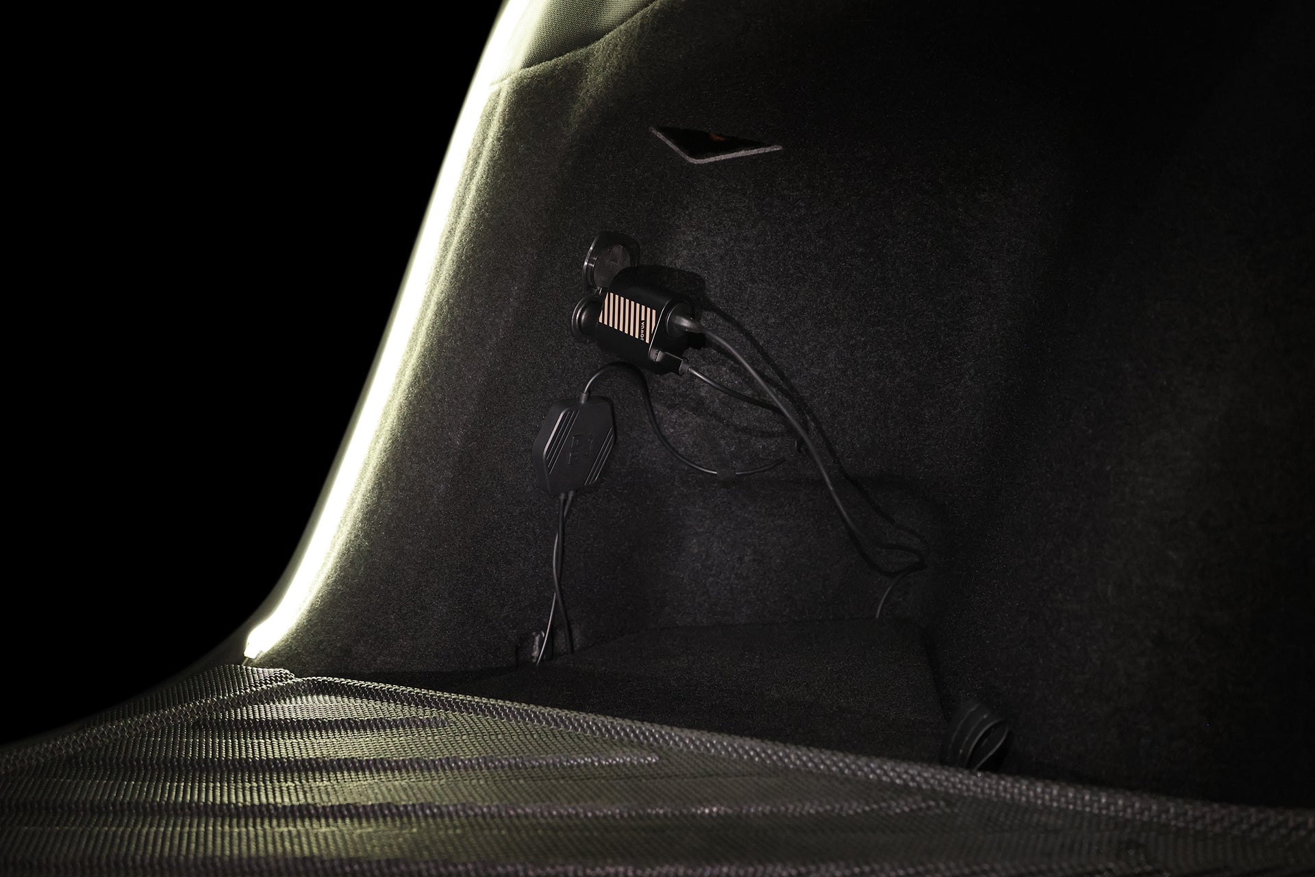 Close-up of a Tesla Model Y trunk interior with the JOWUA Teslussy Light > Type-C Ambient Light & 3 in 1 Car Charger connected and glowing softly, wires plugged into the power outlet against a dark background.