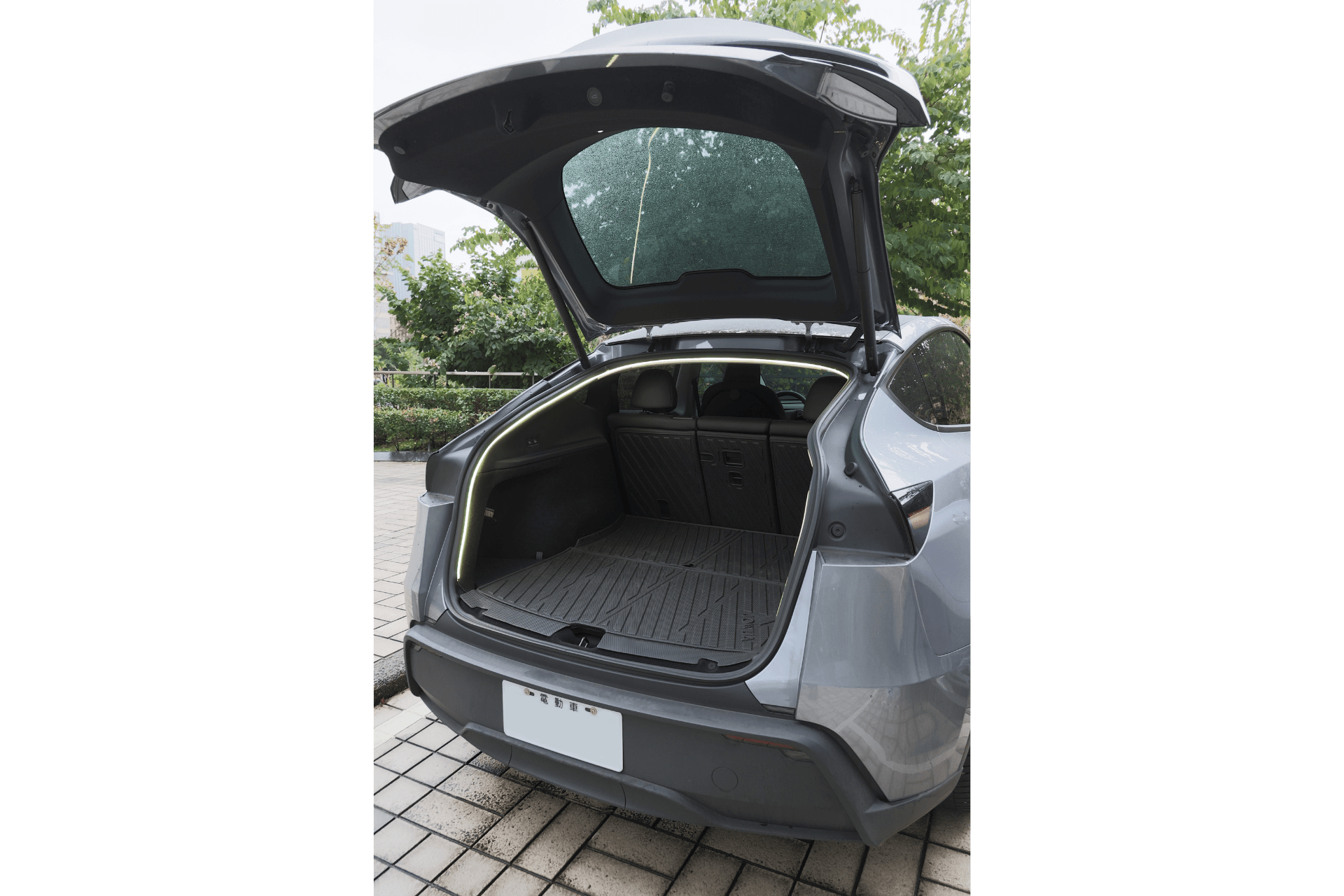 A silver SUV parked on a tiled driveway with greenery behind showcases its empty cargo area, featuring the JOWUA Tesla Model Y Trunk Ambient Light—a USB-C LED strip—illuminating the space.