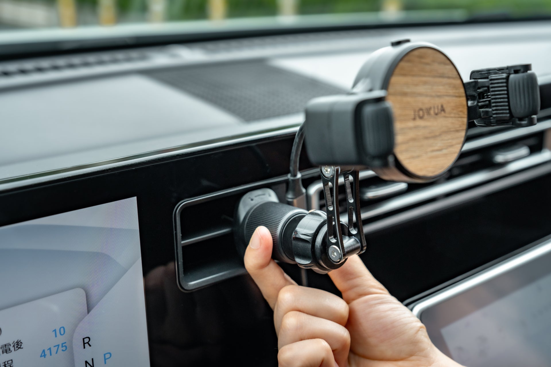 A hand plugs the JOWUA Universal Air Vent Car Mount (Qi2 Universal Roller Mount) into a car’s power outlet, with a phone holder nearby and a digital display screen in the background.