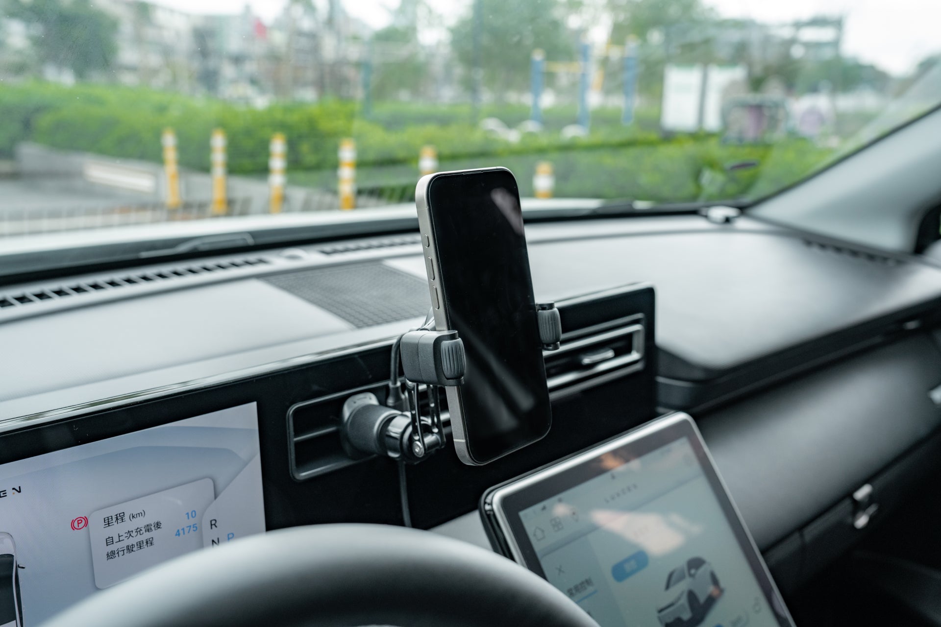 A smartphone is secured to the dashboard using the JOWUA Universal Air Vent Car Mount (Qi2 Universal Roller Mount), positioned beside two digital car displays, with greenery and a road visible outside the windshield.