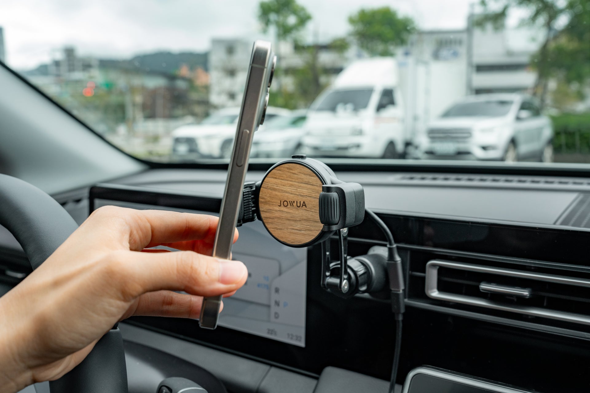 A hand places a smartphone onto the JOWUA Universal Air Vent Car Mount (Qi2 Universal Roller Mount) with a wooden pattern, attached to a dashboard. Parked vehicles and trees are visible outside. Qi2 wireless charging offers added convenience.