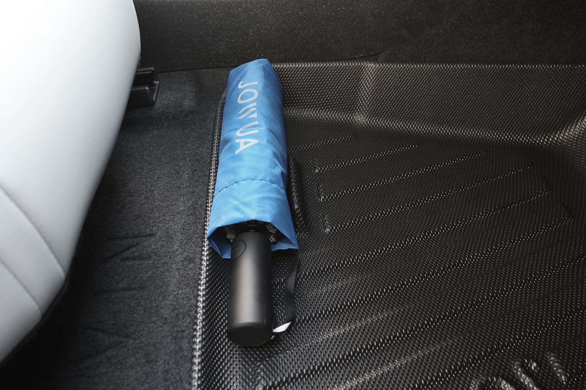 A closed blue umbrella with "JOWUA" printed on its cover rests on a textured black JOWUA All Weather Floor Liner for Volvo EX30, next to a white seat.
