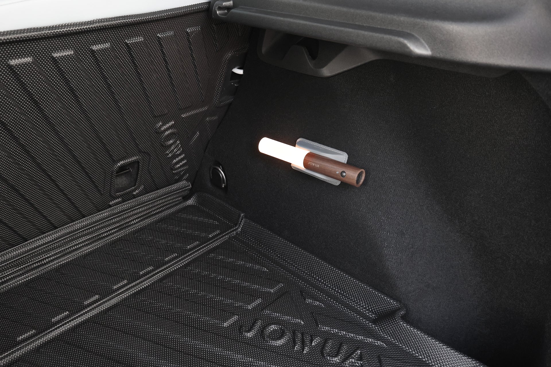 A close-up of a Volvo EX30 trunk shows a JOWUA All Weather Trunk Liner with Rear Seat Cover, featuring custom fit protection beneath a portable flashlight with a brown handle attached to the side panel.
