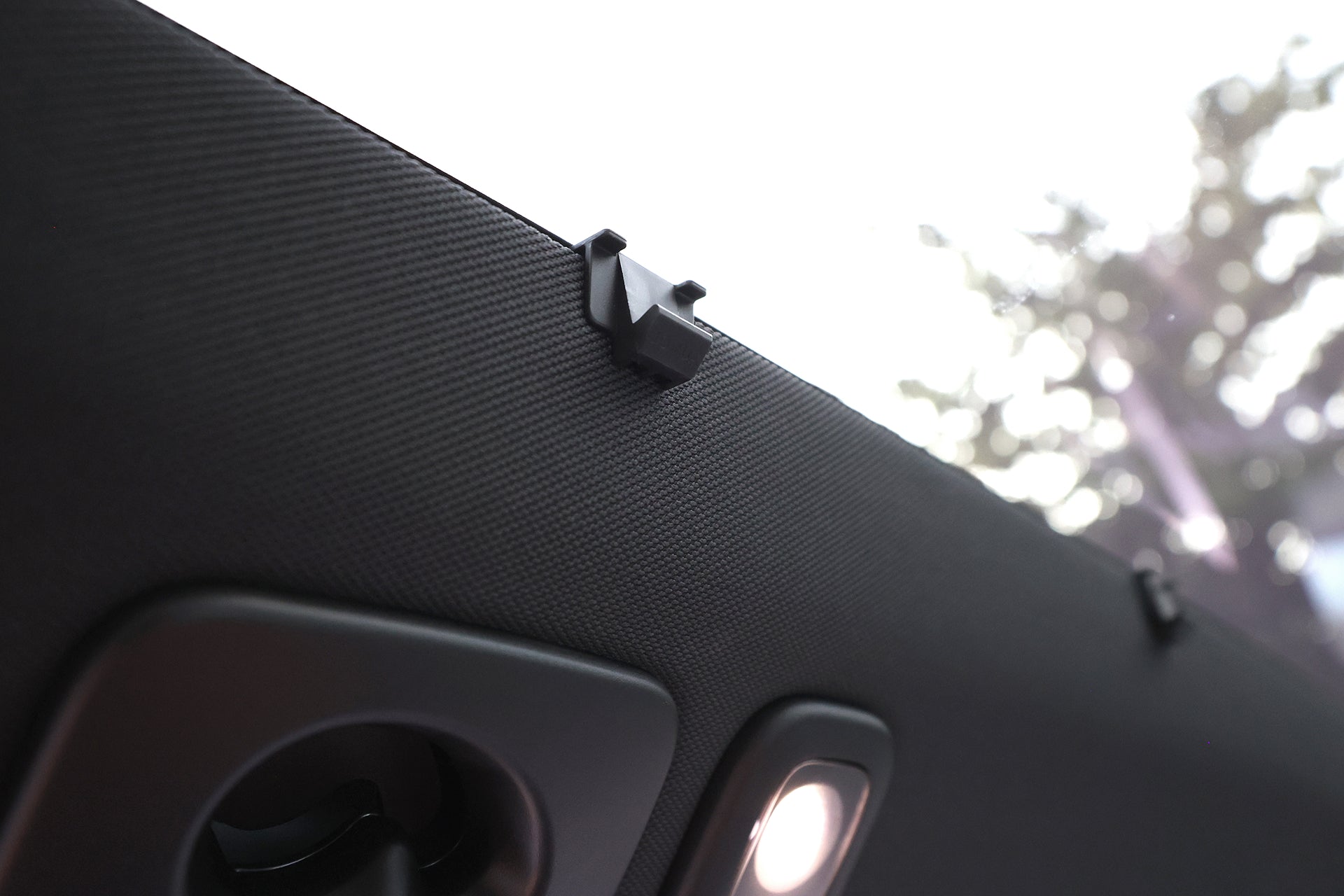 Close-up of the Volvo EX30’s interior roof, showing textured black mesh and built-in lights. Through the window, a JOWUA Glass Roof Sunshade provides added UV protection, with blurry trees visible outside.