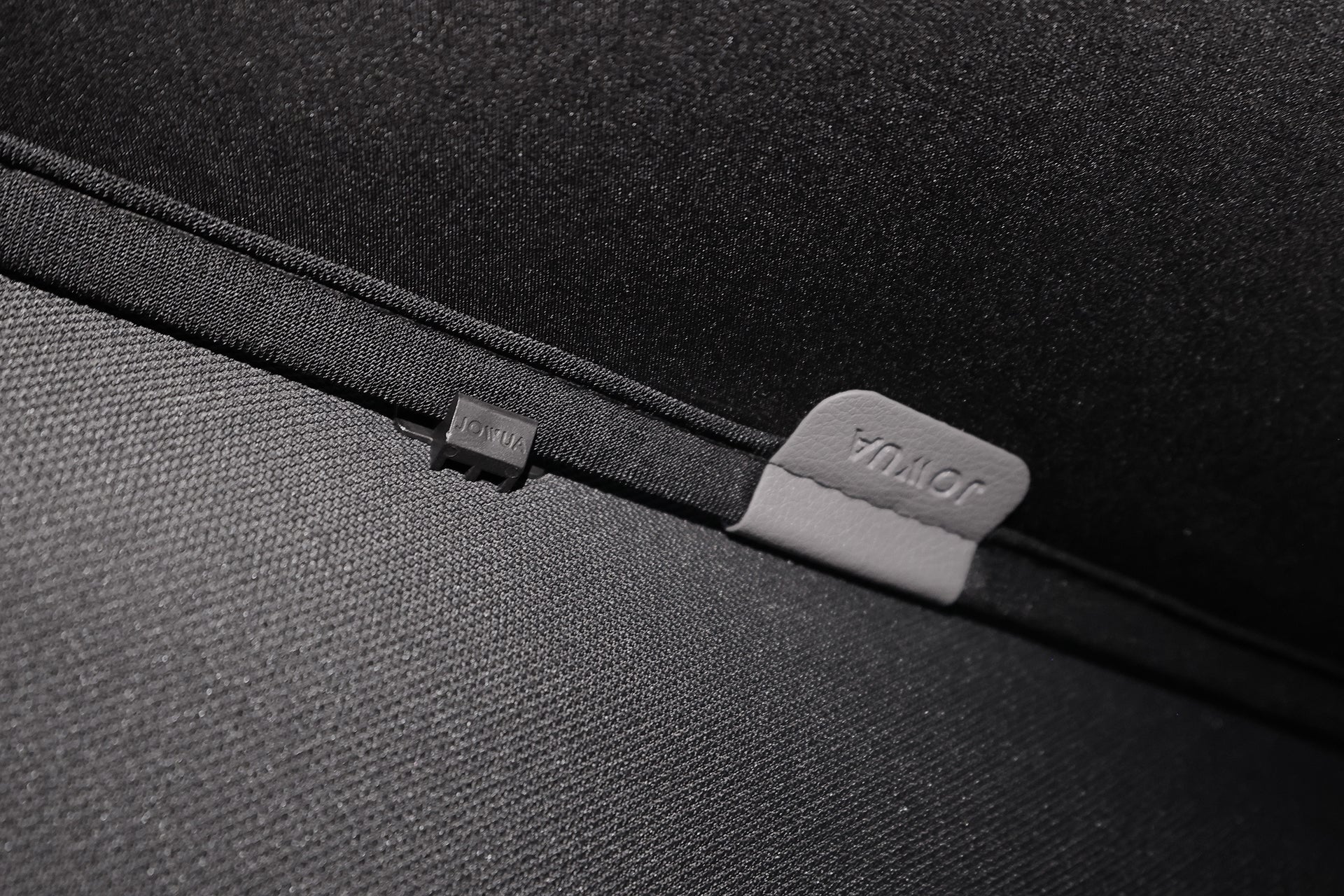 Close-up of a textured black mesh fabric surface with two tags—one small black tag labeled "LOTUS" and a larger gray tag labeled "JOWUA"—featured on the Glass Roof Sunshade for Volvo EX30, designed for UV protection.