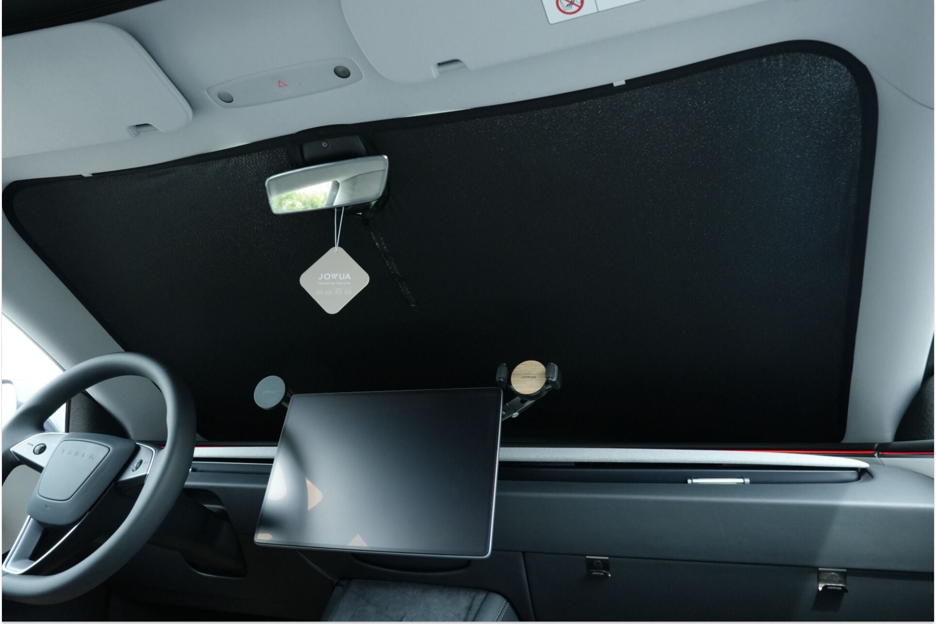 The interior of a Tesla Model 3 includes a JOWUA Windshield Sunshade for all Model 3 versions (M3 / M3HL / M3HL Performance), offering UV protection. It also features a steering wheel, a rearview mirror with a hanging air freshener, and a digital display on the dashboard.