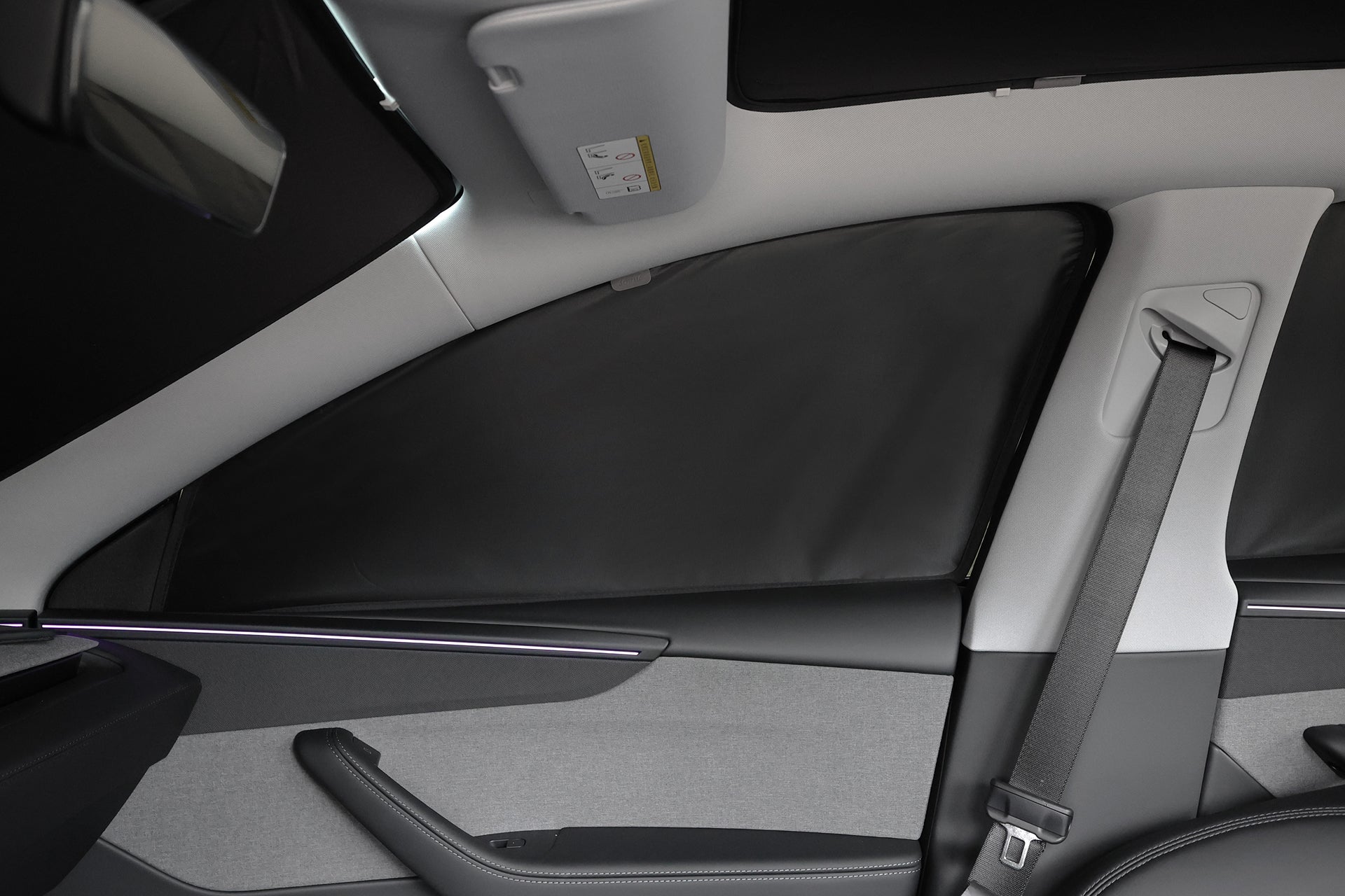 Interior view of a car's rear passenger side featuring the JOWUA Side + Rear Liftgate Window Sunshade for Model Y Juniper (2025+), offering privacy. Visible are the seatbelt, grey and black door trim, and a side airbag label on the roof lining.