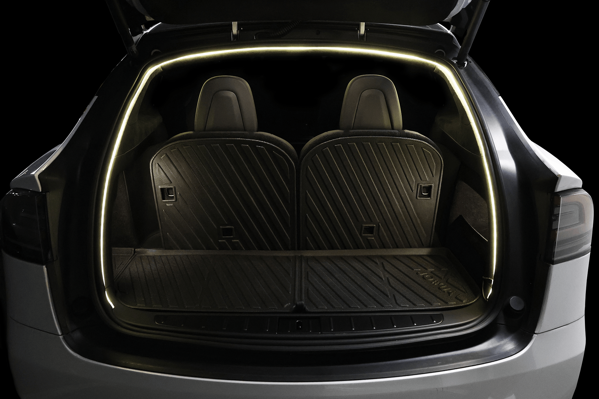 The trunk of a Tesla Model X with rear seats upright is lit by the JOWUA Tesla Model X Trunk Ambient Light - USB-C LED Strip with Magnetic Induction, displaying black mats that cover the trunk floor and seat backs.