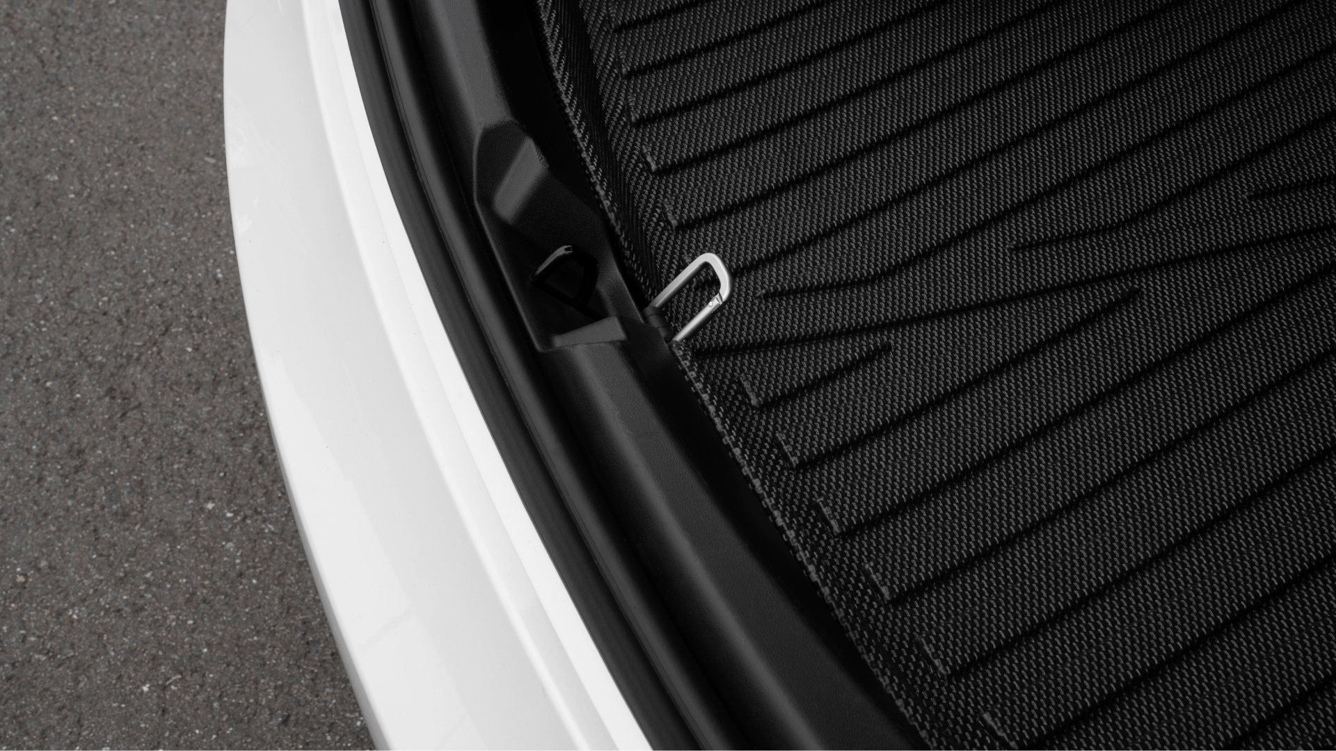 Close-up of a Tesla Model S trunk area displays a JOWUA Snap Hook Tesla Model S/3, along with a black mat featuring a textured pattern. The hook suggests easy access to the sub-trunk compartment. The image captures part of the car's white exterior and the grey pavement below, facilitating effortless opening and closing.