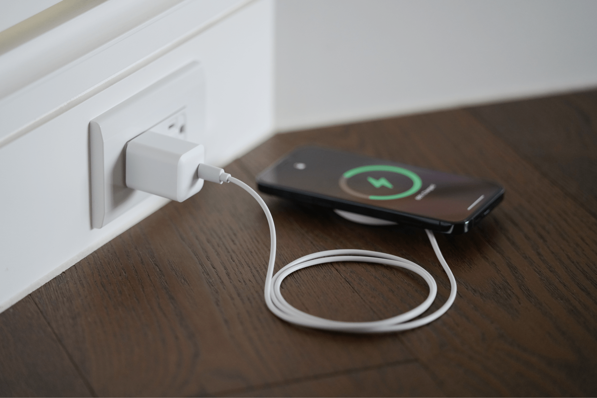 A smartphone rests on a wooden floor, linked to a white USB cable in a wall socket. Its screen shows a green circle with a charging icon, signaling high-speed charging readiness like the JOWUA 100W Portable Air Compressor + Charging Solution Kit handles your favorite Tesla accessories.