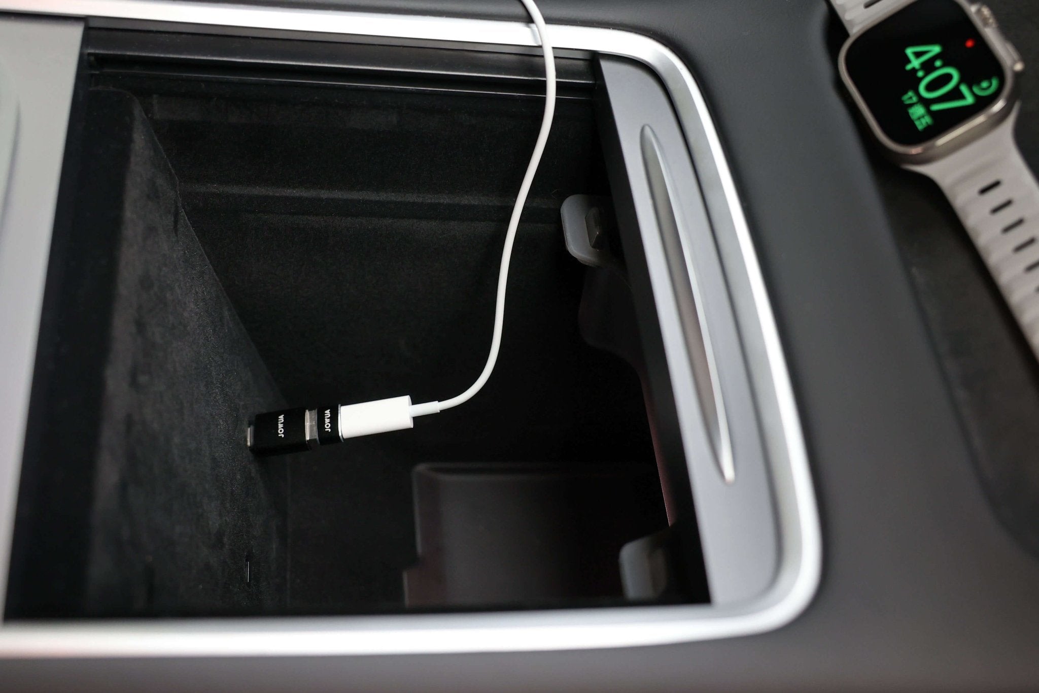 A JOWUA Apple Watch Tesla Charging Kit is plugged into a car’s center console, with a smartwatch showing 4:07 nearby—showcasing convenient on-the-go Apple Watch charging and Power Delivery.