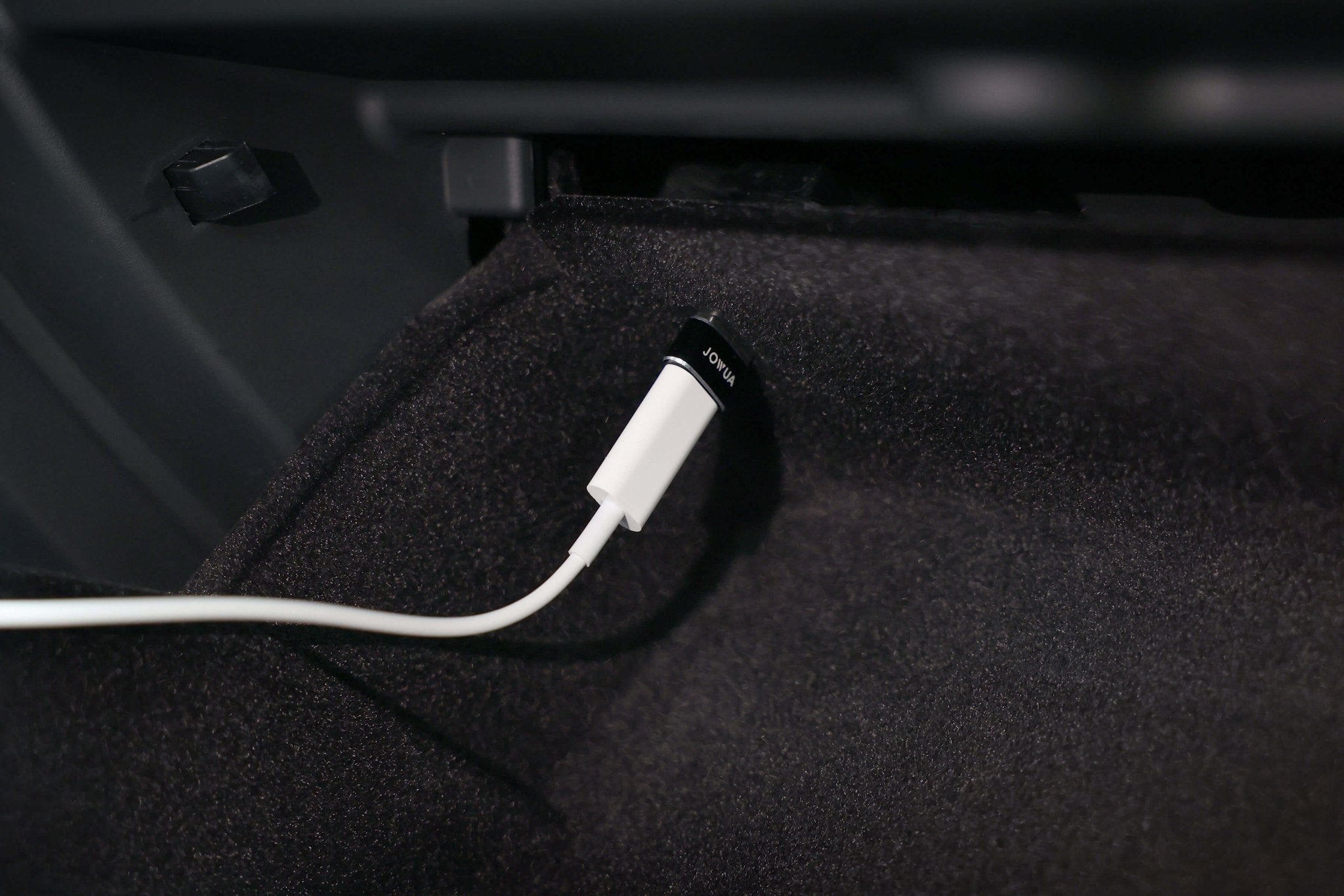 A close-up of the JOWUA Apple Watch Tesla Charging Kit's white USB-C to USB-A adapter cable plugged into a black USB port inside a car’s glove compartment, surrounded by dark interior fabric.