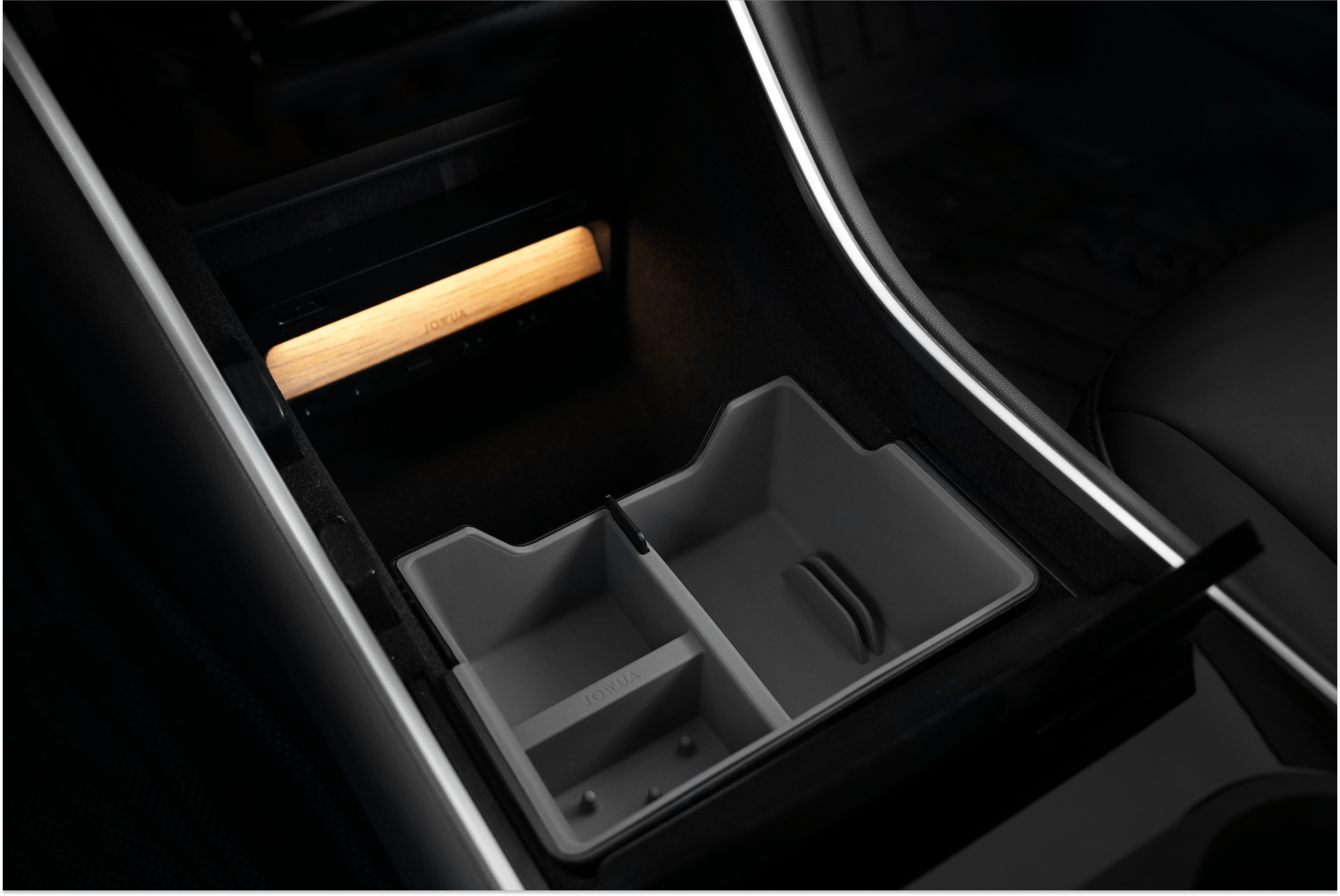The JOWUA Center Console Tray Organizer, designed for pre-refresh Tesla Model 3 and Y vehicles, features an illuminated compartment highlighting a light woodgrain accent. This accessory, crafted from premium hybrid material, includes a plastic organizer with two storage sections. Its sleek design harmonizes with the dark interior surroundings to enhance the soft ambient glow.