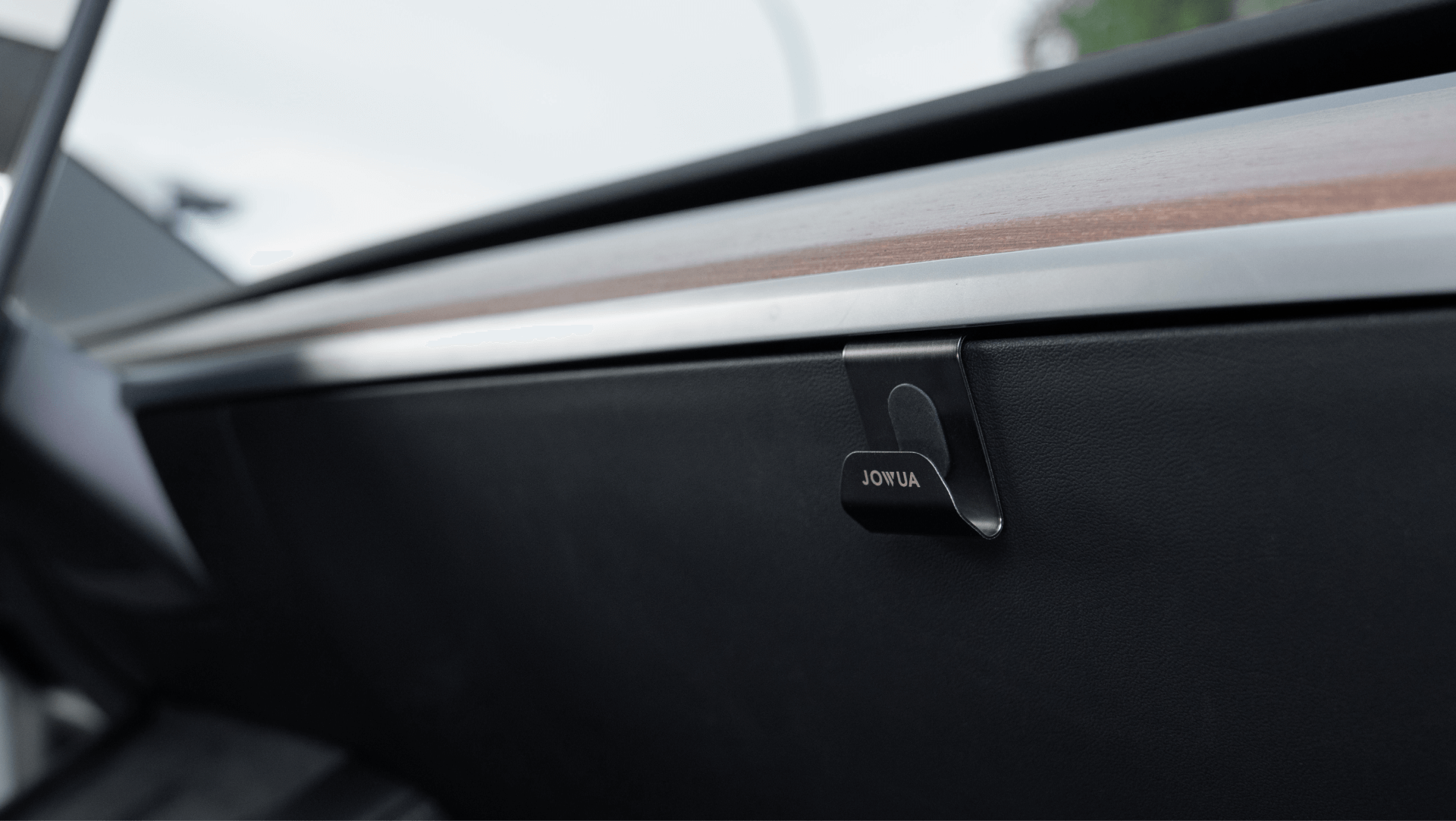 Close-up view of a sleek Tesla Model 3 interior, highlighting a black panel with a small clip labeled "JOWUA." The panel features a wooden trim and metallic edge, enhancing its modern minimalist design. A Glovebox Hook by JOWUA adds functionality to the stylish setting.