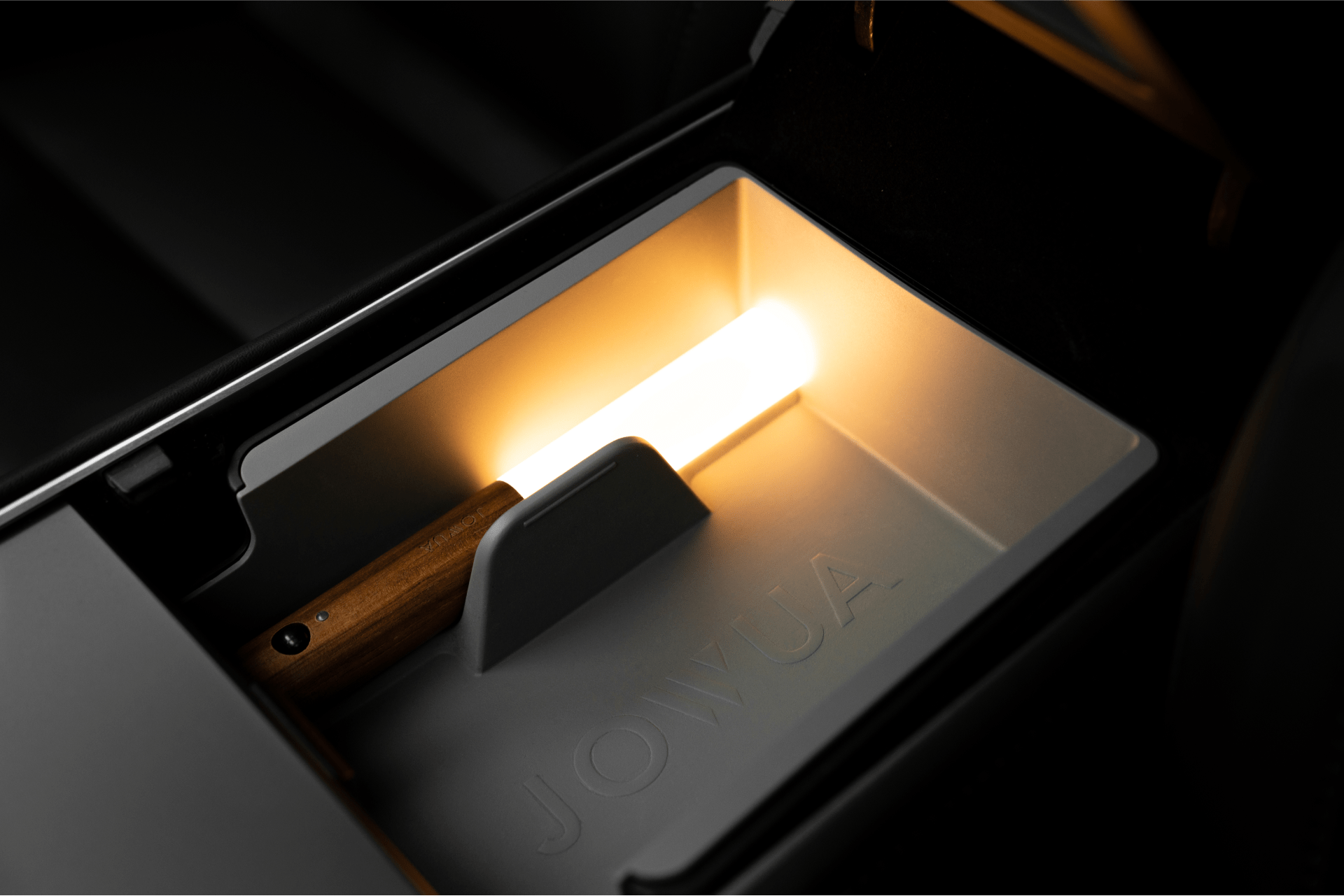 A LED Light Bar designed for the Tesla center console by JOWUA is elegantly displayed in a sleek, illuminated tray with the brand's embossing. The warm light emphasizes its minimalist design, echoing the sophistication of Tesla Interior Lighting.
