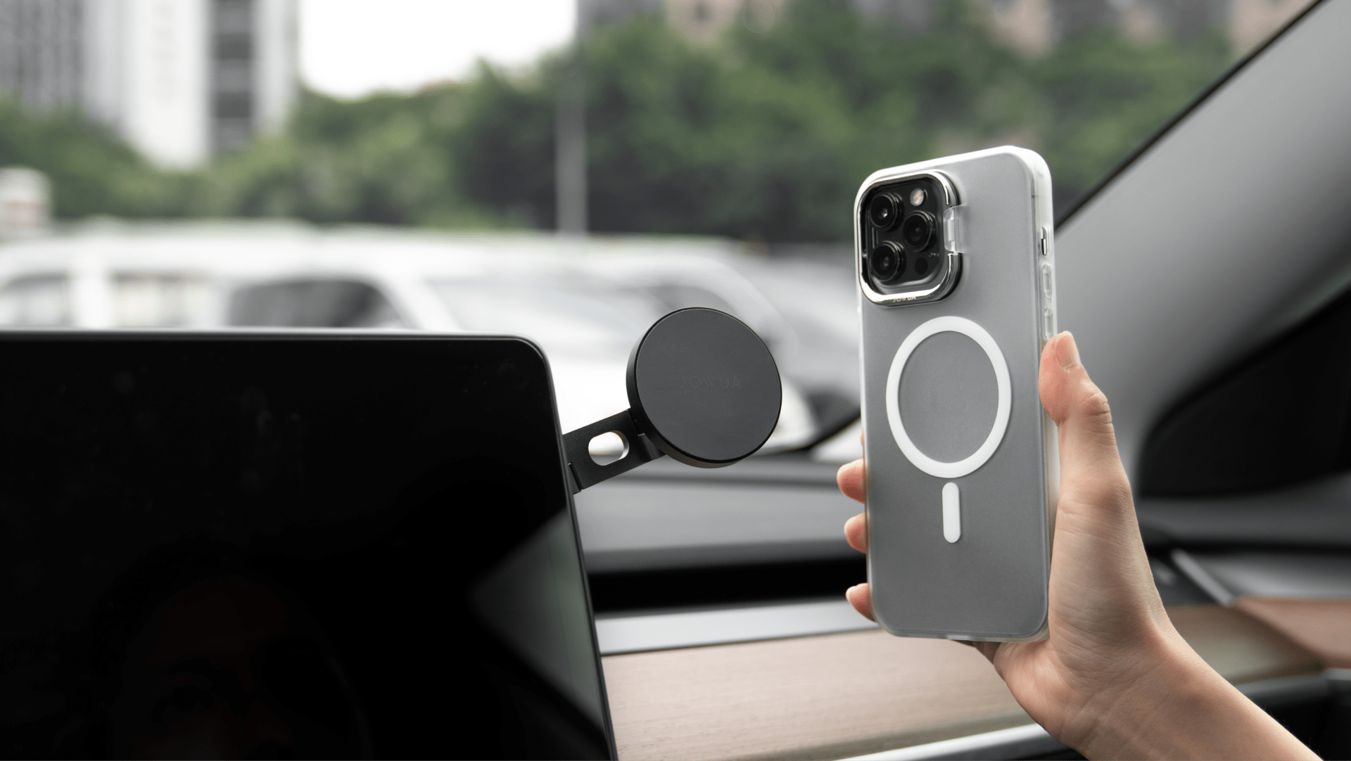 A person holding an iPhone fitted with the JOWUA iPhone Case for Tesla Car Mount, featuring MagSafe capabilities and a circular design on the back, near a magnetic car mount. The background shows blurred trees and buildings outside a car window.