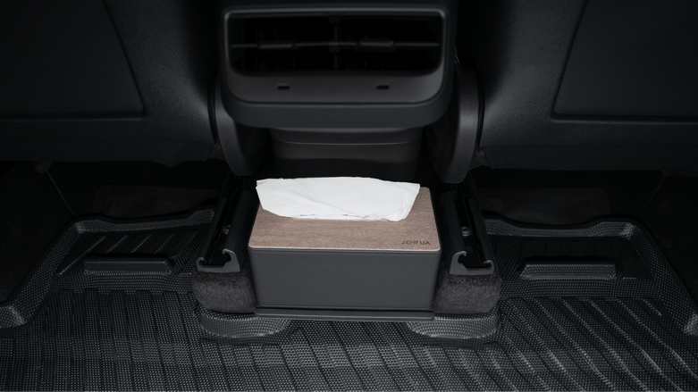 The JOWUA Premium Interior Essentials Set (RHD) – Model Y Juniper (2025+) box of tissues with a wooden lid is placed under the rear air vents, resting on the rubber floor mat beneath the car seat in this right hand drive vehicle.