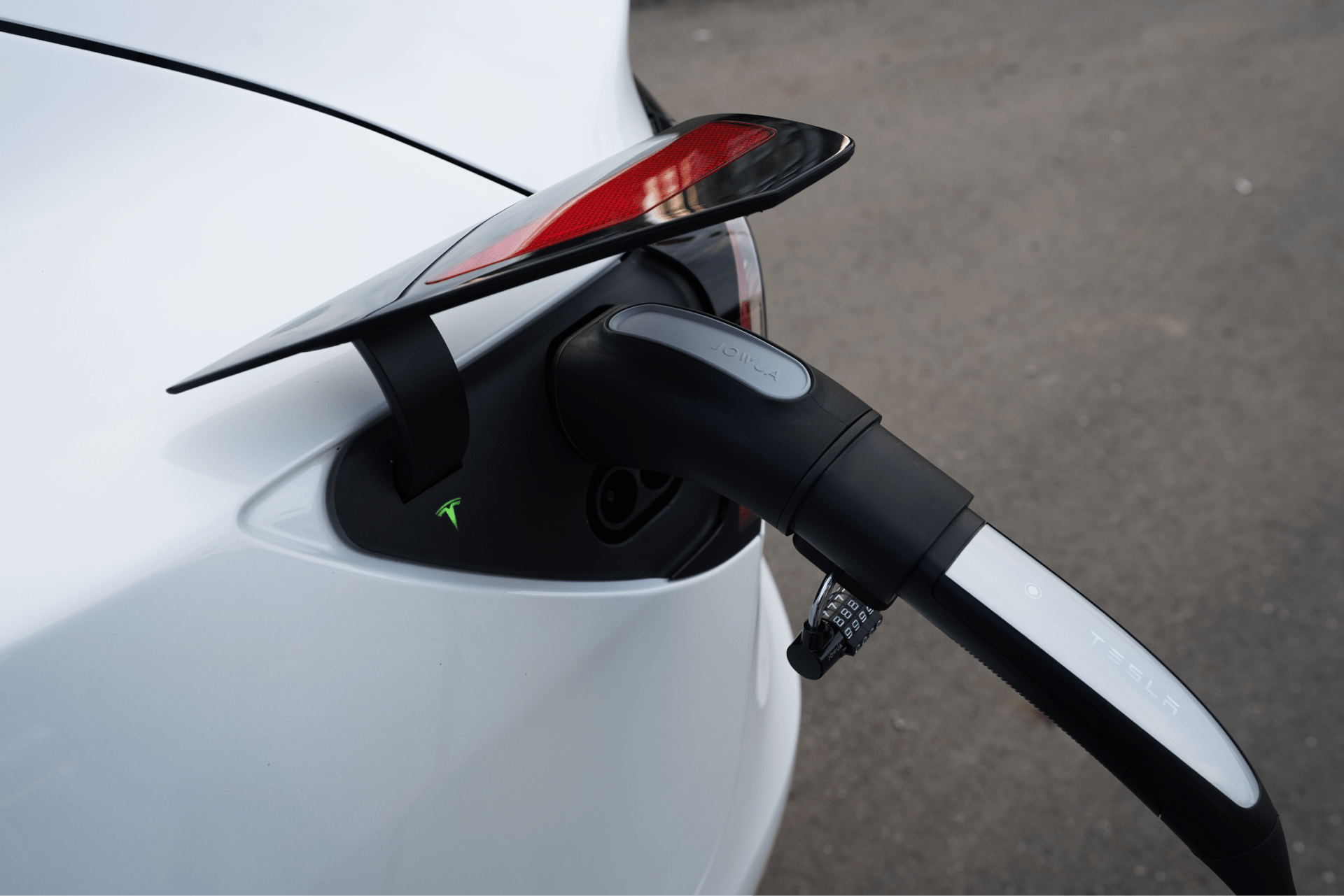 A close-up view captures a white electric car being charged, featuring the JOWUA TPC(NACS) to CCS2-AC (TYPE2) Adapter connected to the vehicle's port. The tail light and part of the rear can be seen against a blurred asphalt backdrop, accentuating the sleek padlock-inspired design.
