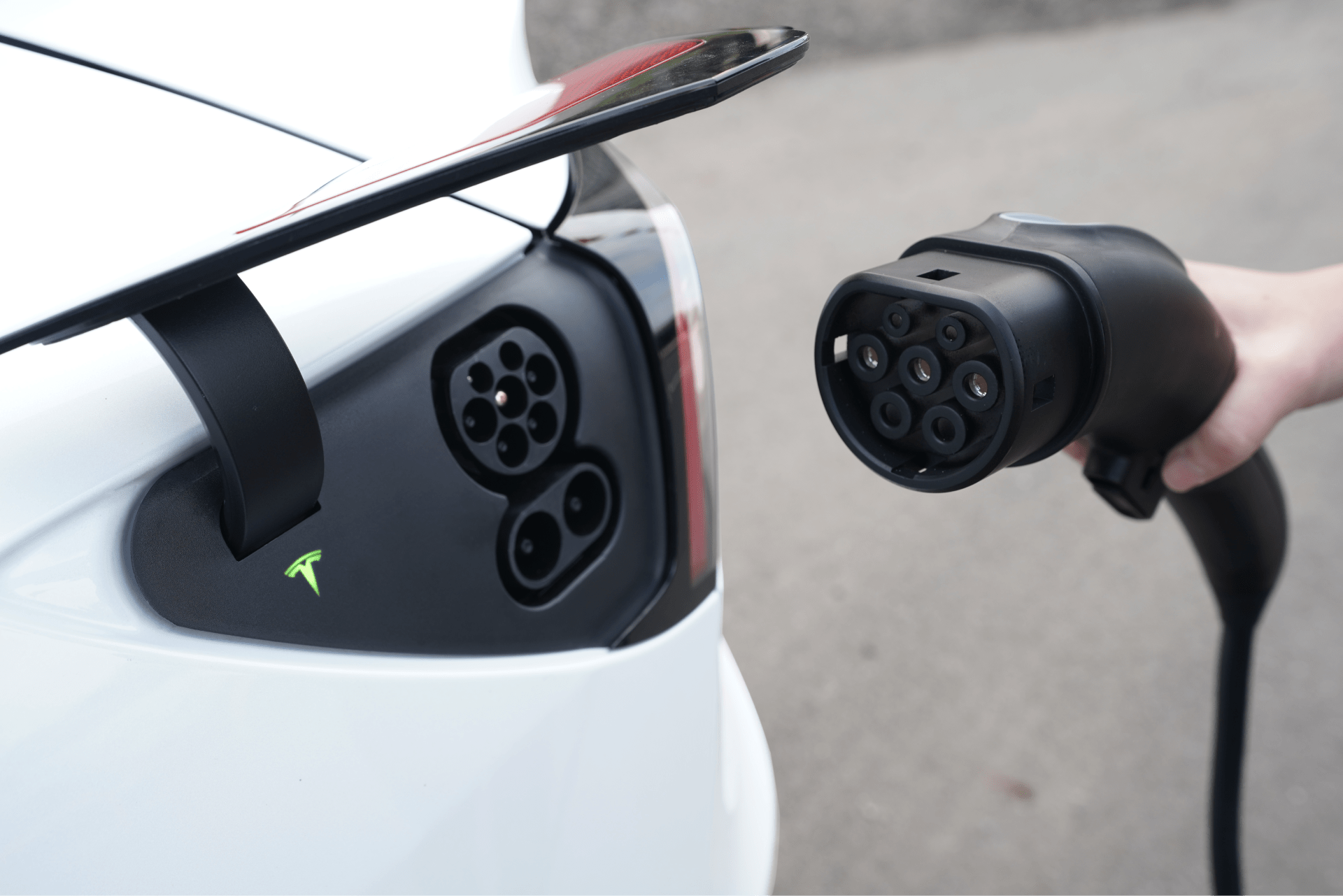 A close-up of a white electric car being charged highlights its sleek design. The charging port is open as a hand grips the TPC(NACS) to CCS2-AC (TYPE2) Adapter by JOWUA, showcasing the large, black plug with multiple connectors. Emphasizing connector durability, this adapter adheres to IEC 62196-2 standards for efficient and secure charging.