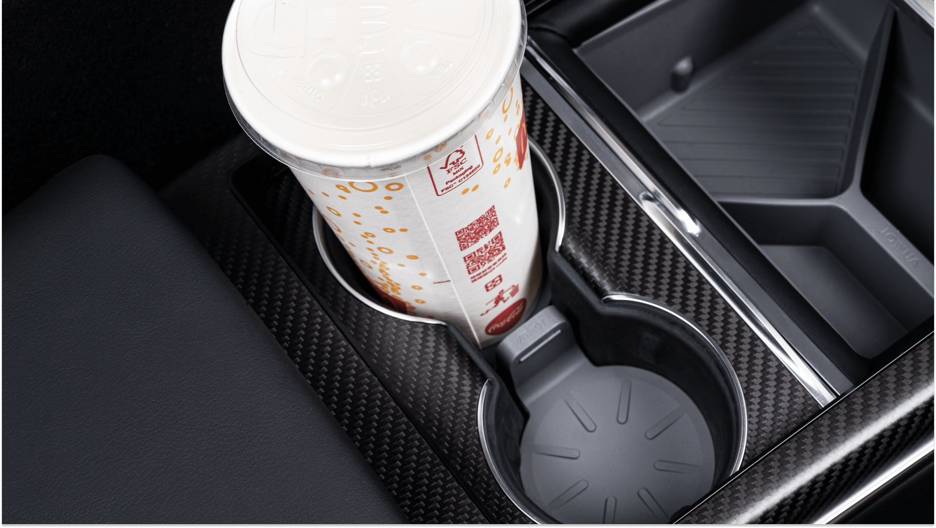 A large soft drink cup with a white lid fits neatly in a car's cup holder beside an empty spot, perfectly highlighted by the sleek carbon fiber console. The JOWUA Center Console Set for Tesla Model S/X ensures that your black leather seat interior remains tidy and stylish.