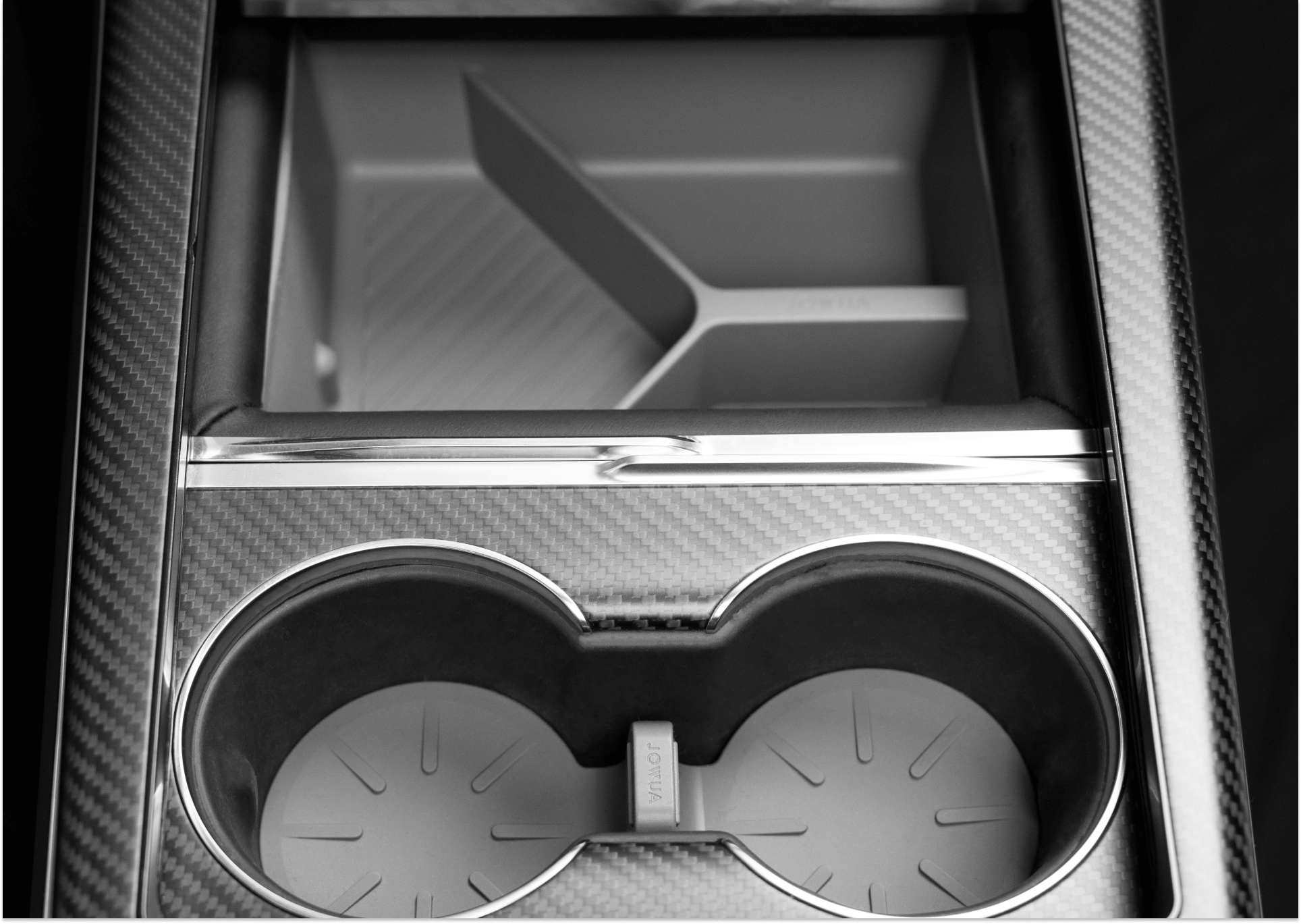 A close-up of the JOWUA Center Console Set for Tesla Model S/X showcases two cup holders equipped with premium silicone coasters labeled "VIGOR." Positioned above the cup holders is a small storage compartment with a divider. The console features a stylish carbon fiber finish, enhancing its modern appeal.