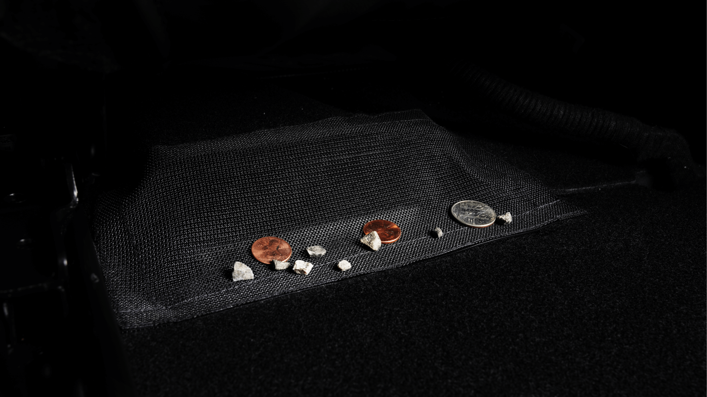 Coins and small debris rest on a black mesh fabric against a dark background, similar to how loose change might accumulate on the all-weather floor liners in the JOWUA Tesla Model Y Juniper (2025+) Essential Bundle for right-hand drive models.