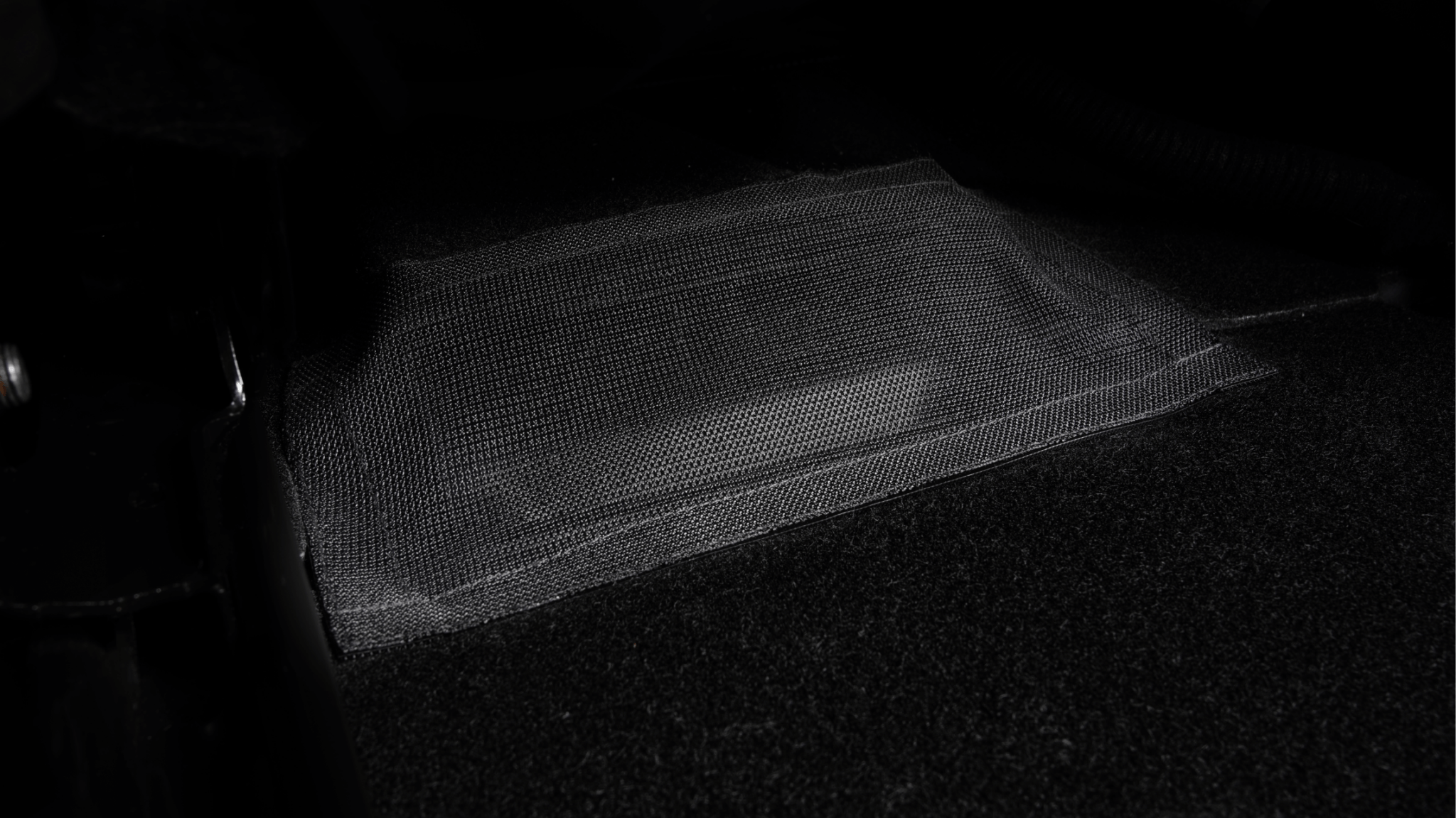 A close-up image of a JOWUA Rear Under Seat Air Vent Cover, showcasing its rectangular patch of water-resistant mesh fabric adhered to a dark surface. The texture and weave are clearly visible, while the surrounding area is shadowed, suggesting low lighting conditions similar to the sleek interior details in a Tesla Model Y.