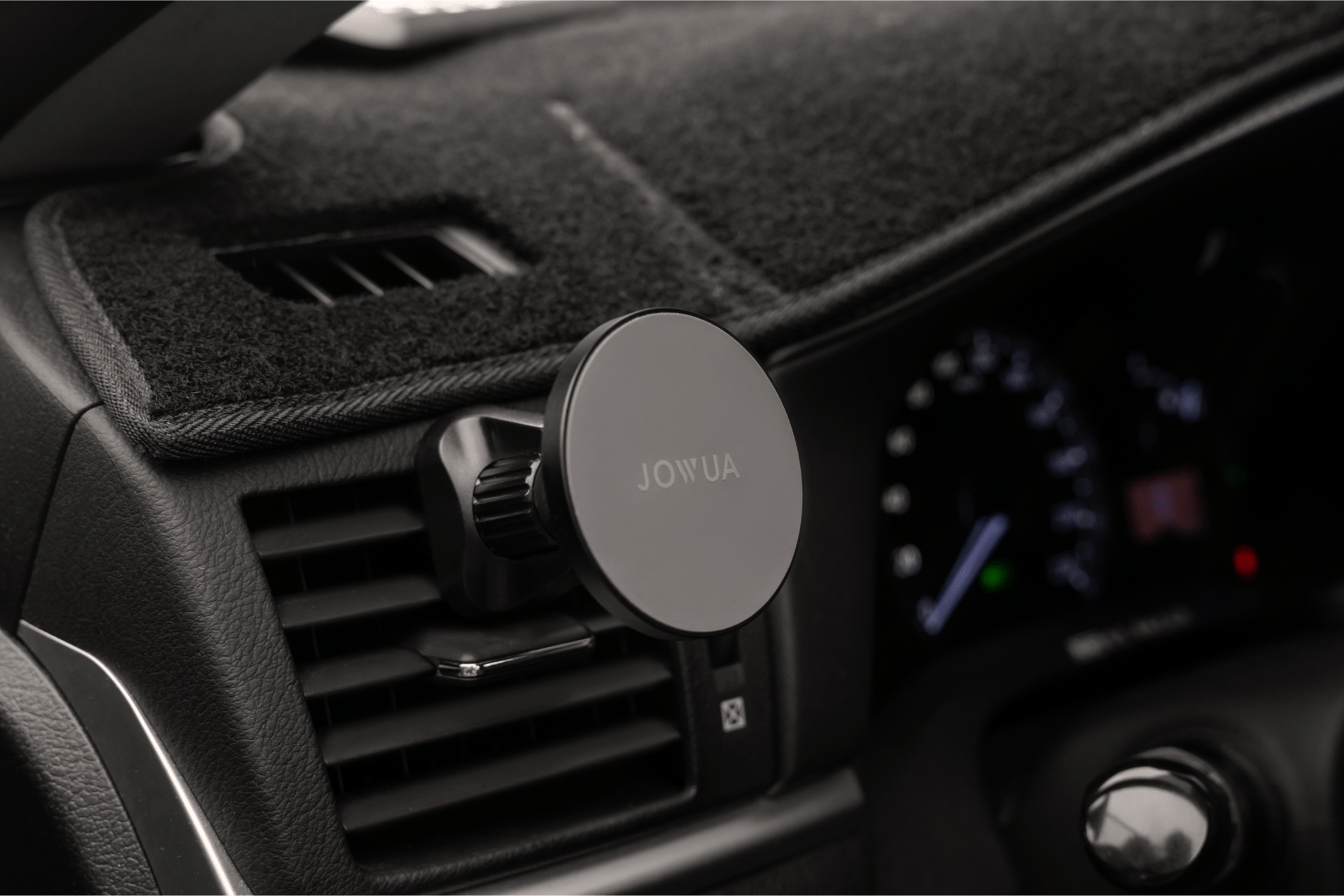 The image showcases a close-up of a car dashboard with the JOWUA Universal MagSafe Car Mount clipped onto an air vent, providing Qi2 Wireless Charging. The speedometer and other dashboard controls are slightly blurred in the background, keeping your device powered while on the move.