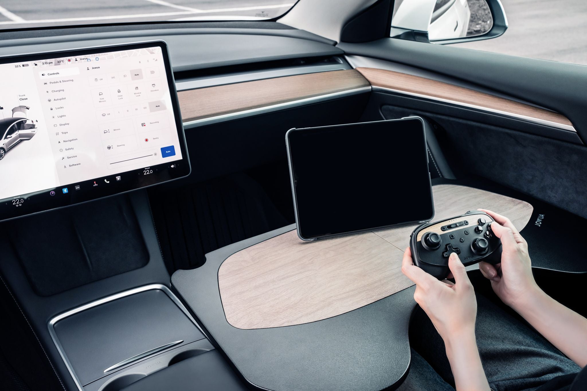 A person sitting in a car interior, using a game controller with a touchscreen and tablet mounted on a wooden dashboard. The car has a sleek, modern design with digital displays.