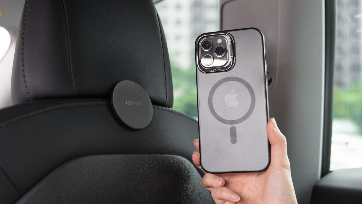 A person holds an iPhone with a MagSafe-compatible case inside a car. The phone is near a magnetic mount attached to the back of a car seat, ready for attachment. Blurred greenery is visible through the car window.