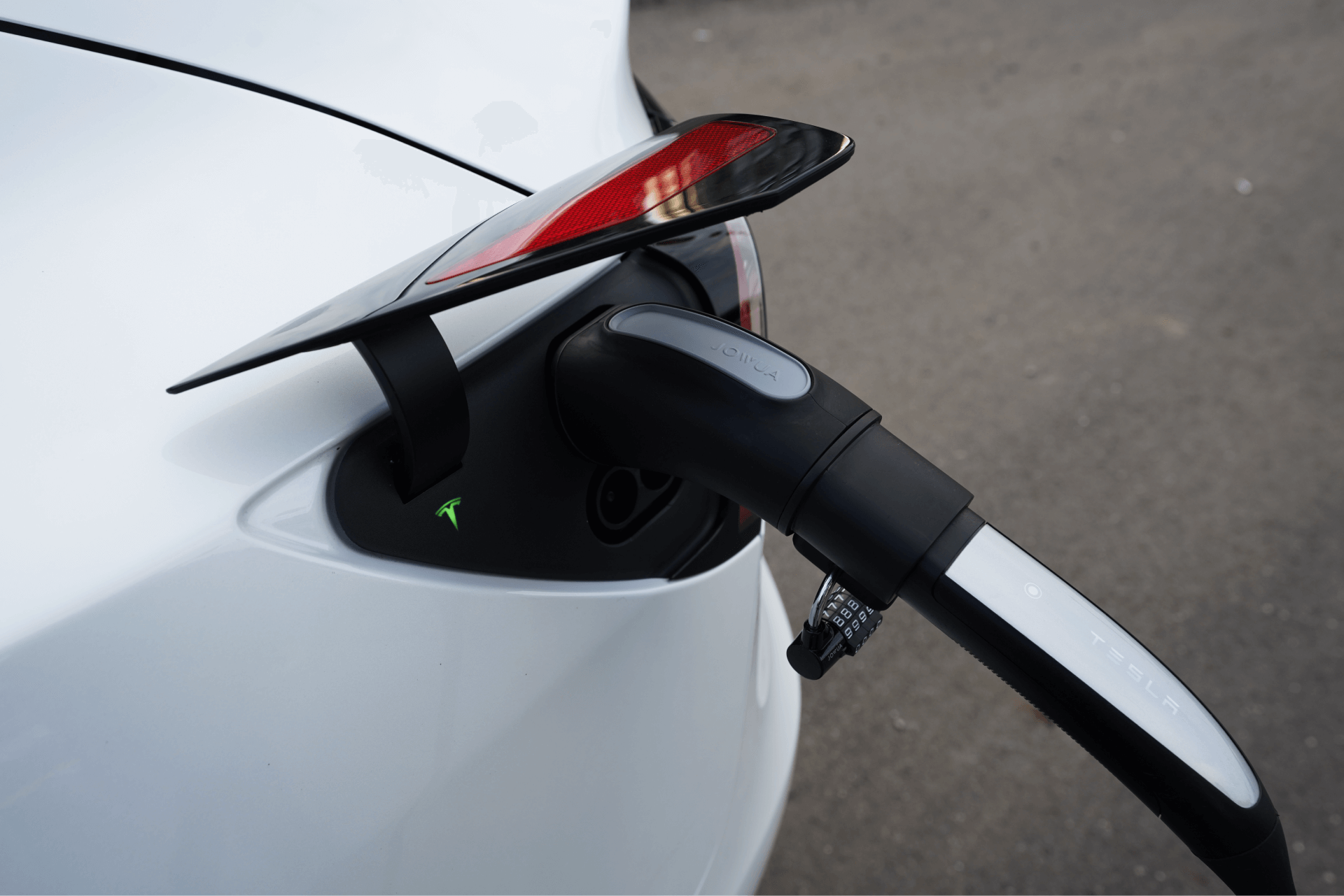 A close-up of a white electric car being charged. The charging cable is plugged into the back of the car, with a logo on the cable, against a background of gray pavement.