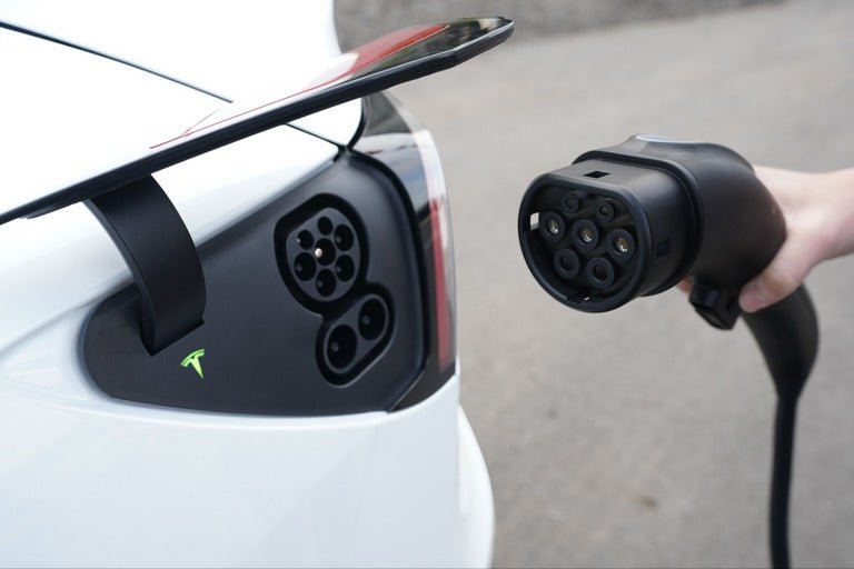 Close-up of a person holding a charging plug near an electric car's charging port. The car is white and features a green logo next to the port, with part of a rear spoiler visible above.