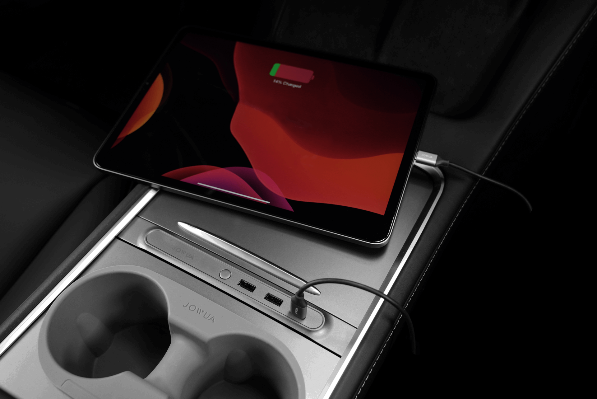A tablet displaying a low battery symbol is charging in a car's central console. The console has cup holders, a closed compartment, and cords connected to USB ports. The tablet screen shows abstract artwork.