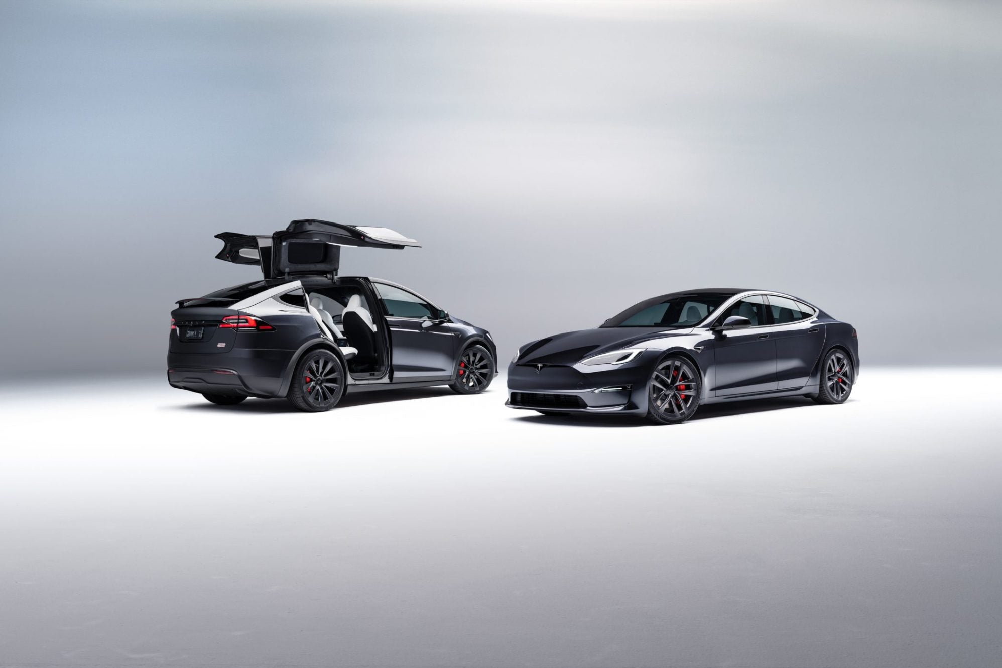 Two sleek black electric cars are parked in a minimalist studio. One has gull-wing doors open, showcasing the interior. Both vehicles have a modern, aerodynamic design against a neutral background.