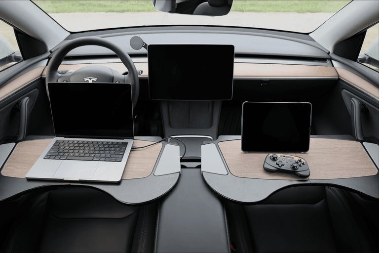 A Tesla car interior with two desks; the left holds a laptop, the right holds a tablet and game controller. Two large screens are mounted on the dashboard, and the steering wheel is visible.