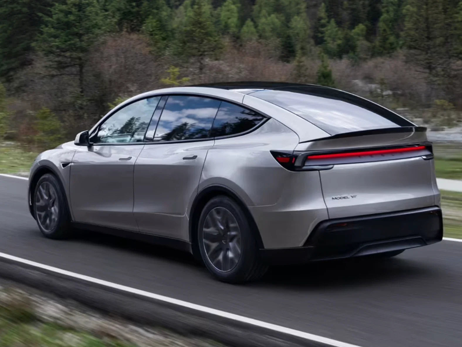 A silver electric SUV drives on a road surrounded by trees and greenery, viewed from the rear side angle.